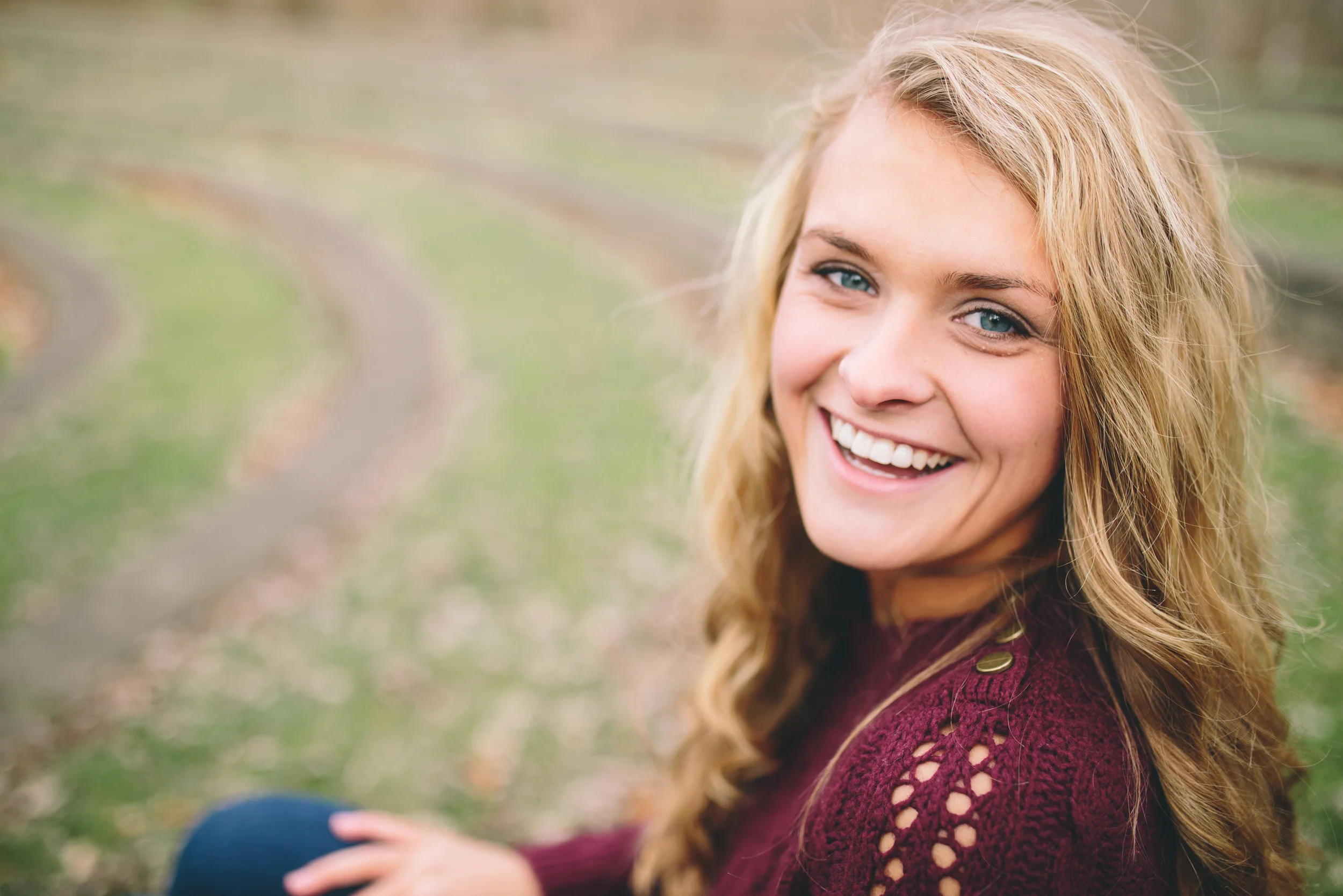 Close-up of a smiling woman with blonde hair and blue eyes, wearing a maroon sweater with cut-out details, sitting outdoors on a grassy path.