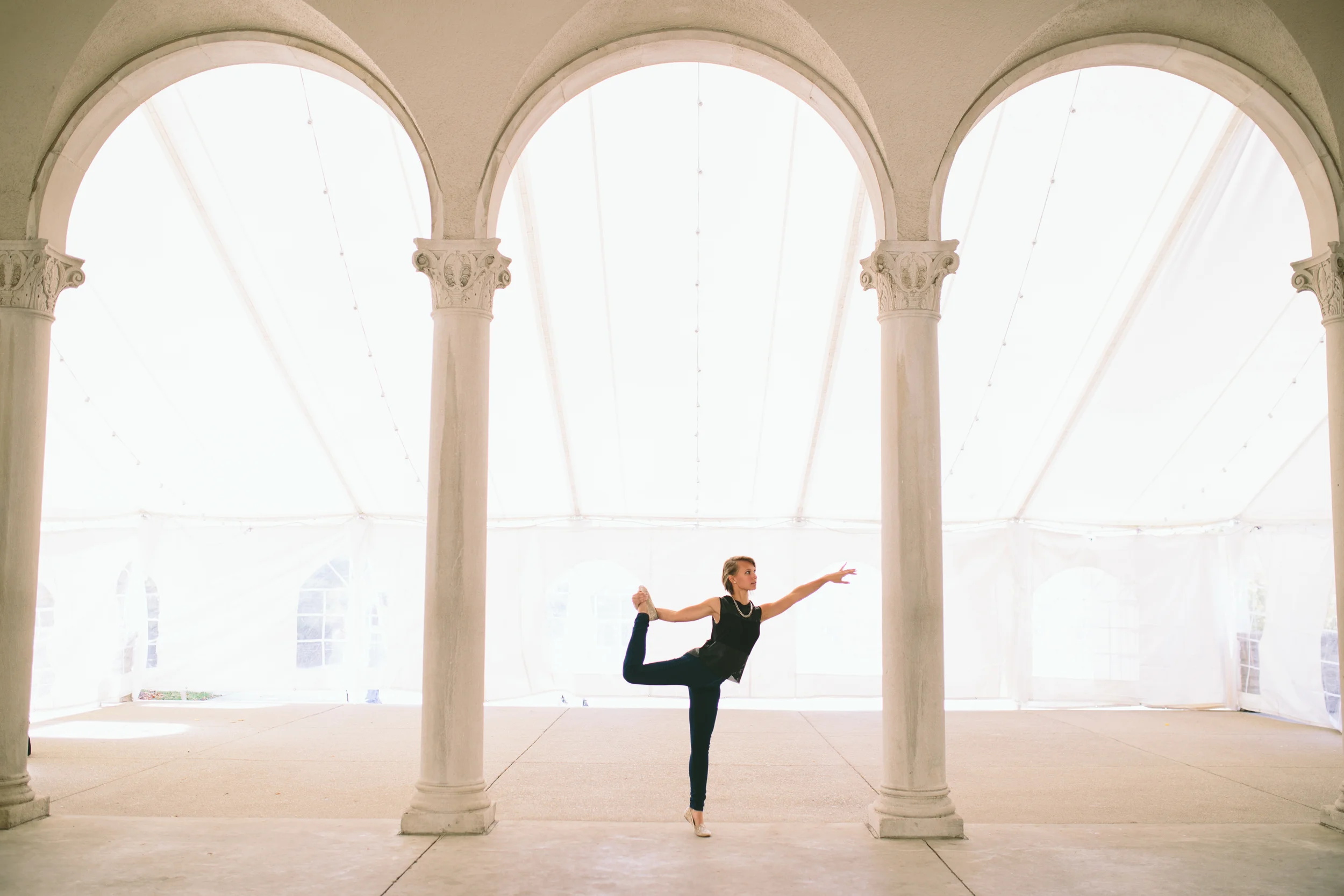 A woman practicing ballet in a large, open hall with tall, arched windows and ornate columns.