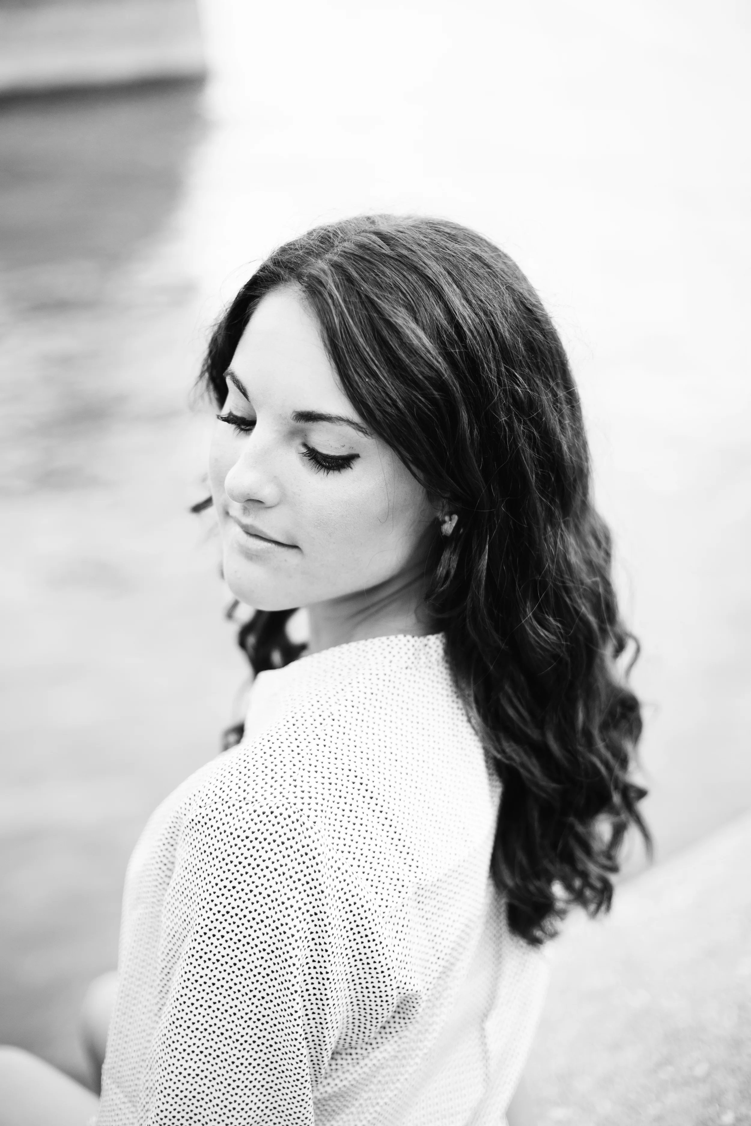 A black and white photo of a young woman with long wavy hair, wearing a textured top, with eyes closed and a calm expression, leaning slightly to her right, against a blurred background.