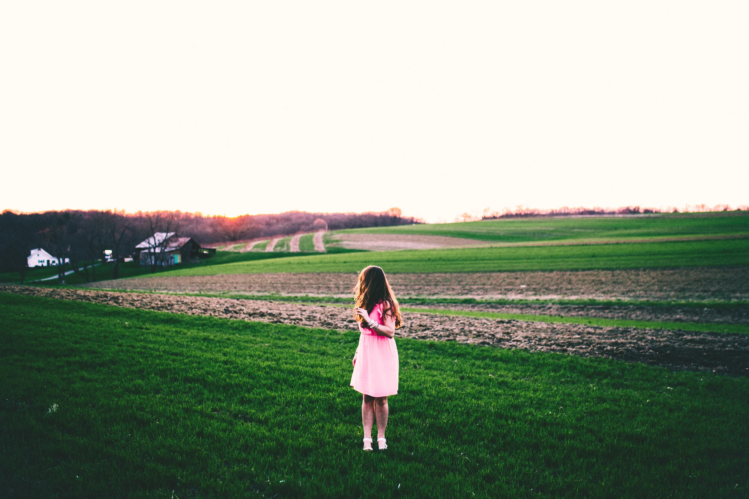 A young girl with long hair wearing a pink dress walking on a grassy field toward an open landscape during sunset.