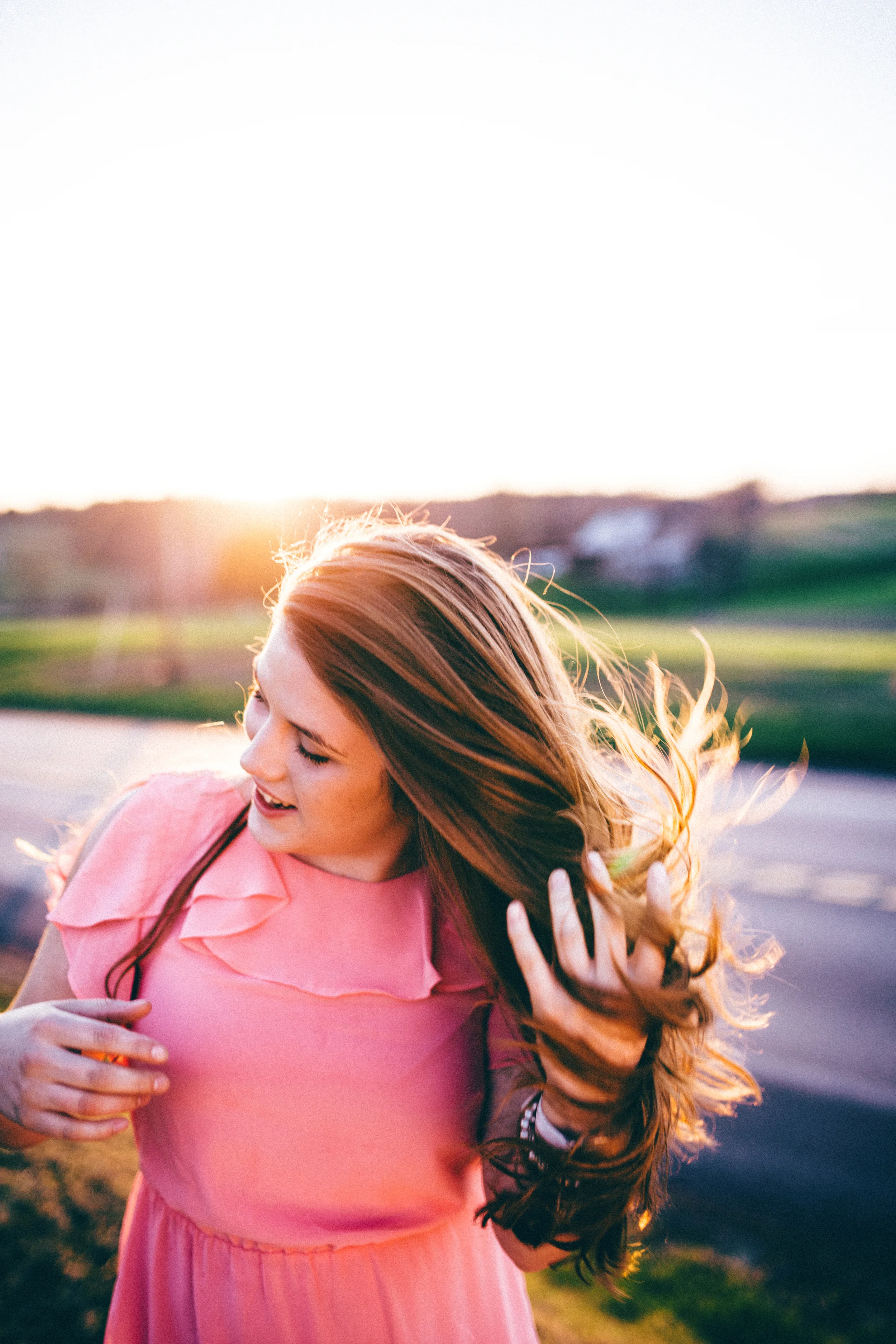 A woman with long, wavy red hair smiling and holding her hair as she walks outdoors during sunset, wearing a pink dress.