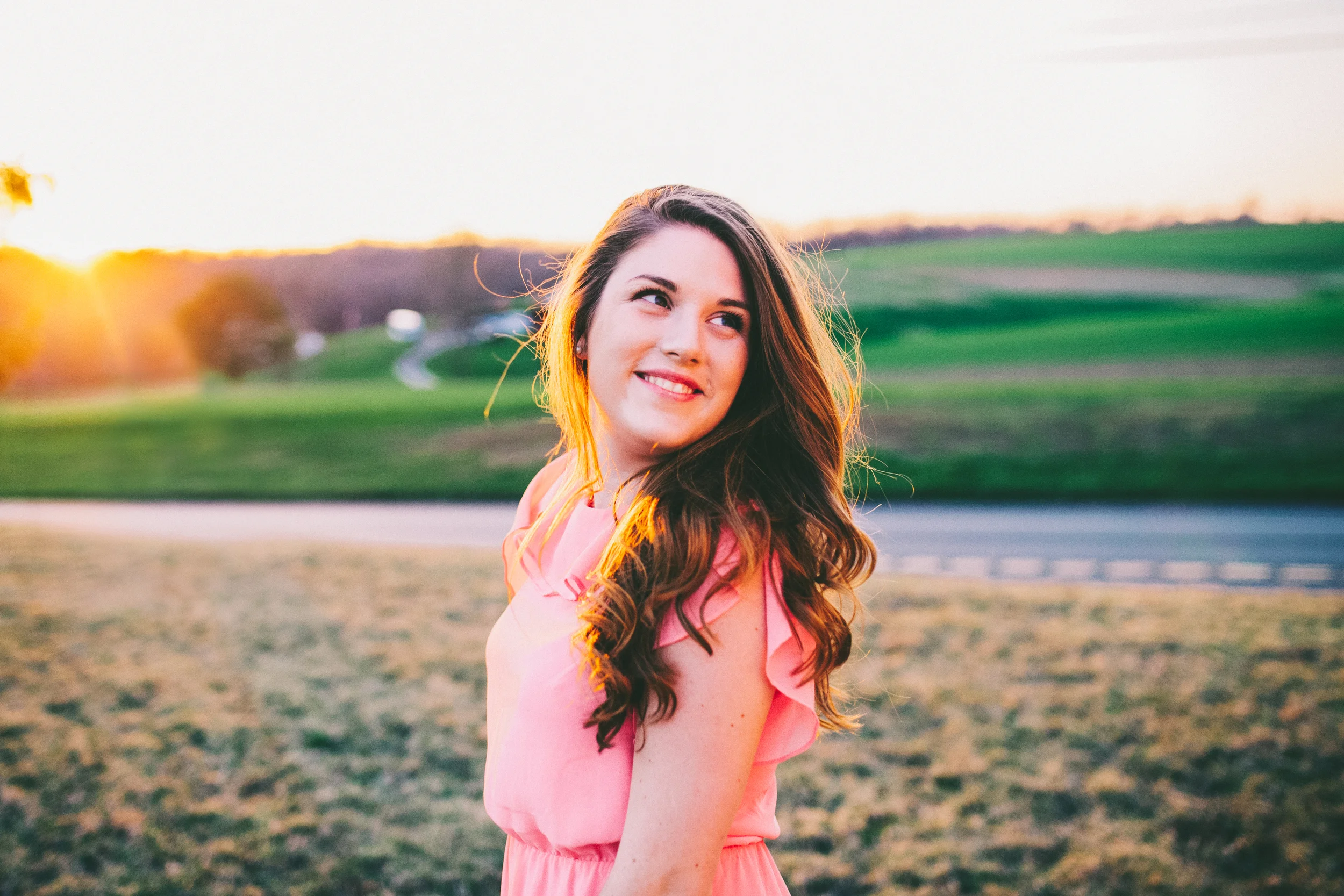 A young woman with long wavy brown hair smiling in a pink dress outdoors at sunset, with a grassy field and distant hills in the background.