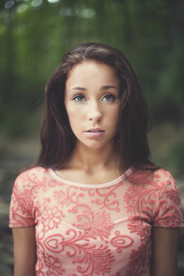A young woman with long brown hair, blue eyes, and light skin standing in a green outdoor setting, wearing a pink lace top with floral embroidery.