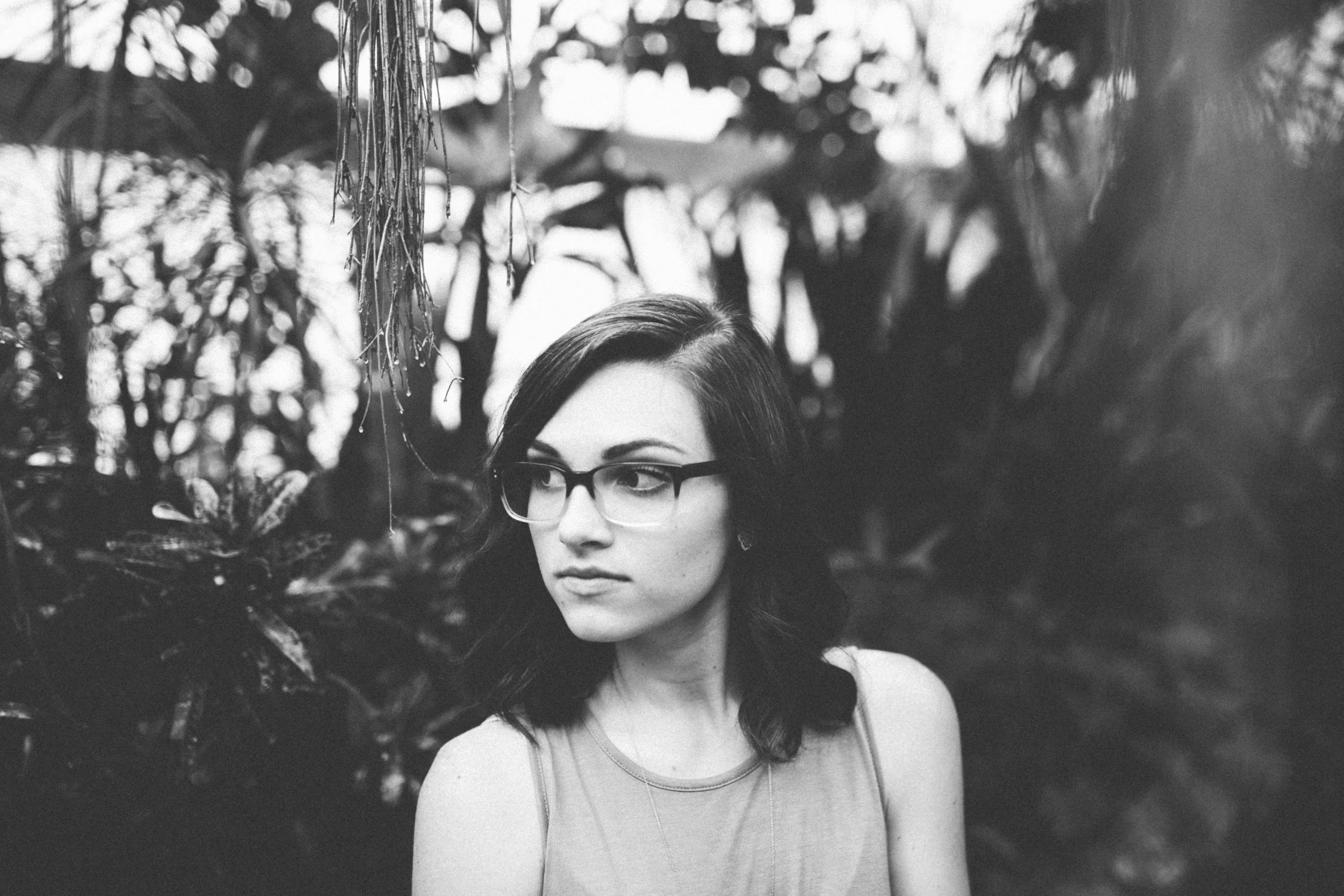 A woman with glasses and dark hair in a garden or forest setting, with foliage and trees in the background, captured in black and white.