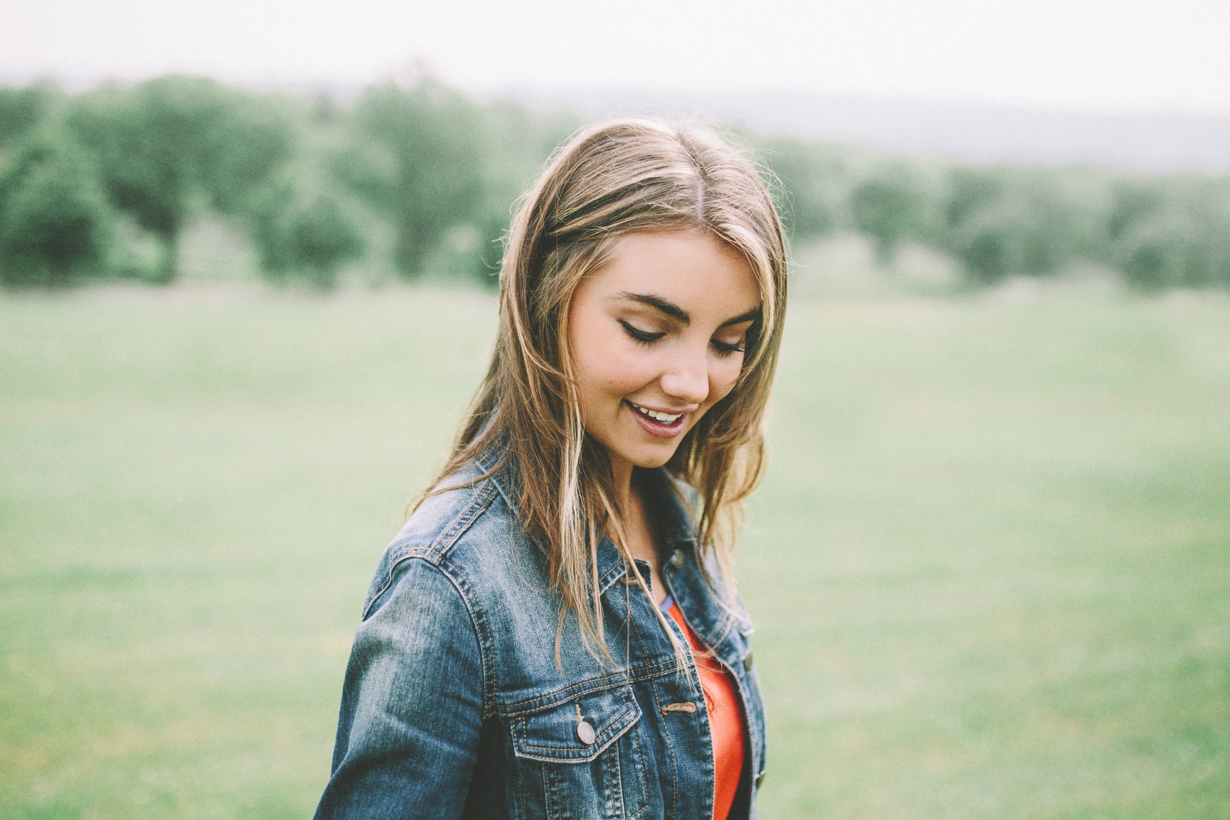 A young woman with long hair smiling outdoors in a green field, wearing a denim jacket and red top.
