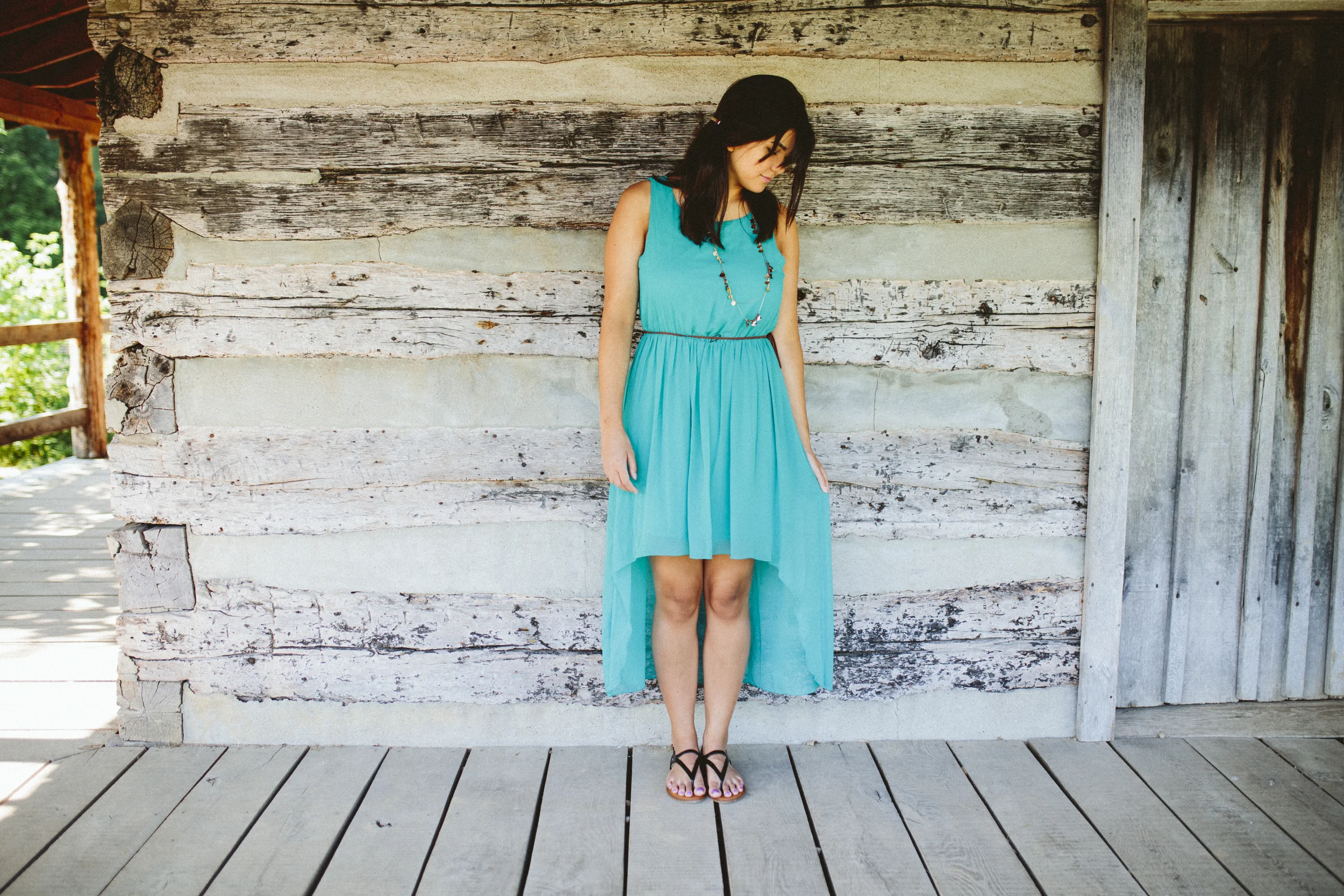 A young woman in a turquoise dress with a high-low hemline standing against a worn wooden wall.