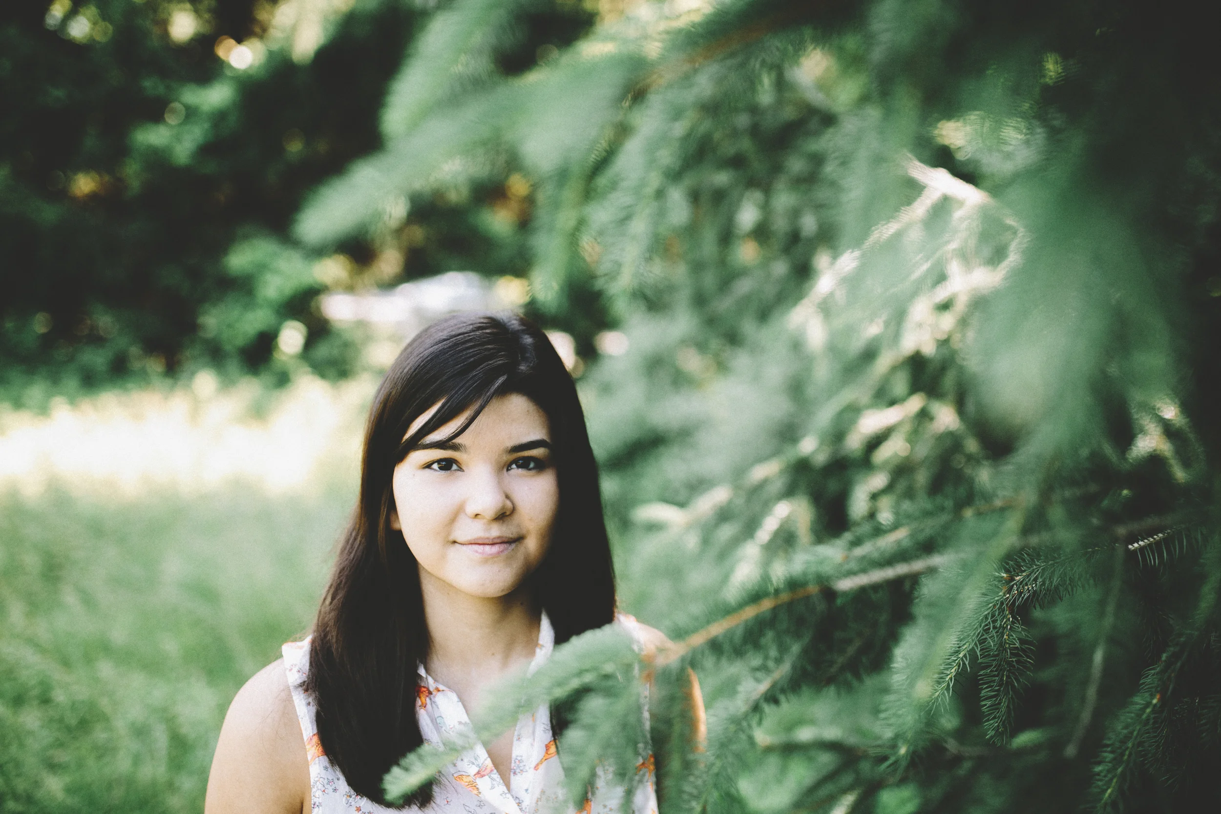 A young woman with long dark hair standing outdoors near evergreen trees, with greenery in the background.