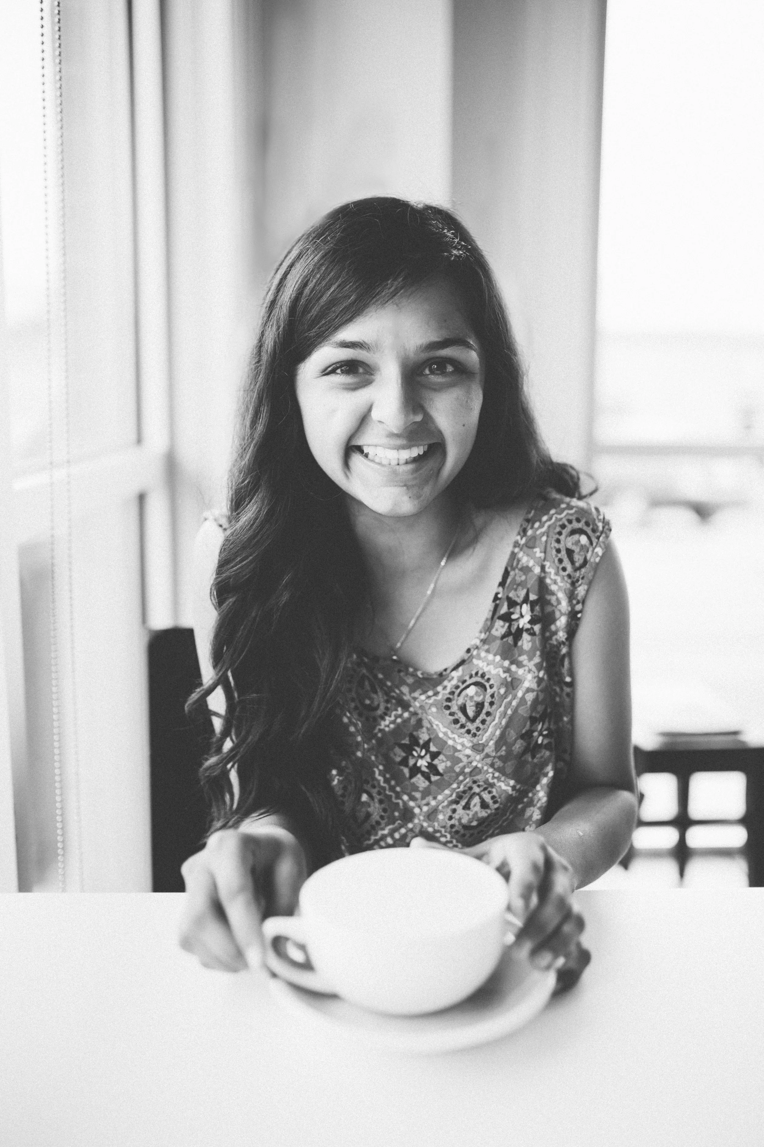 A woman with long, dark hair smiling and holding a coffee mug, sitting at a table indoors next to a window.