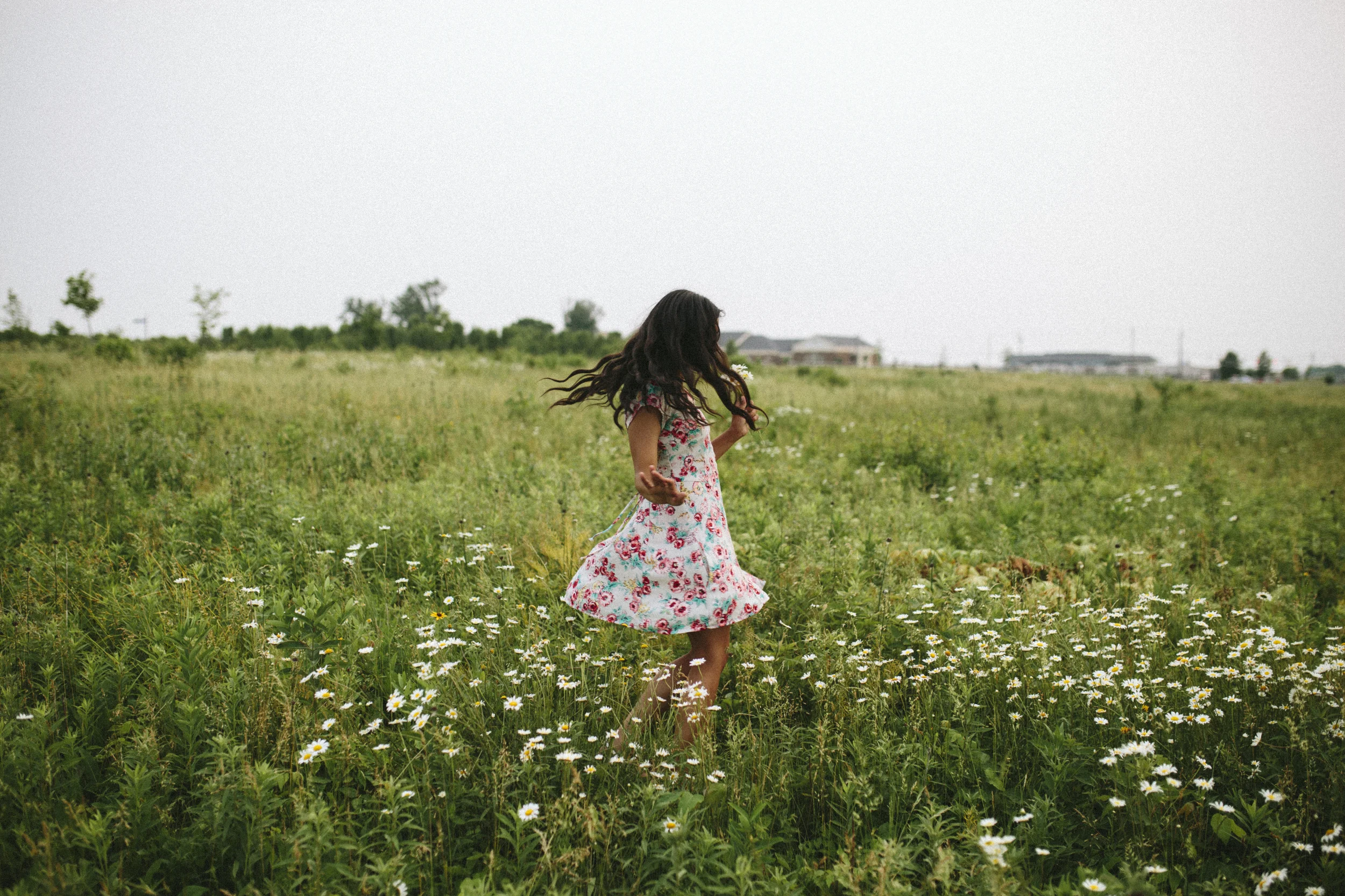 A young girl with long dark hair, wearing a floral dress, is barefoot and walking through a green field filled with small white daisies.