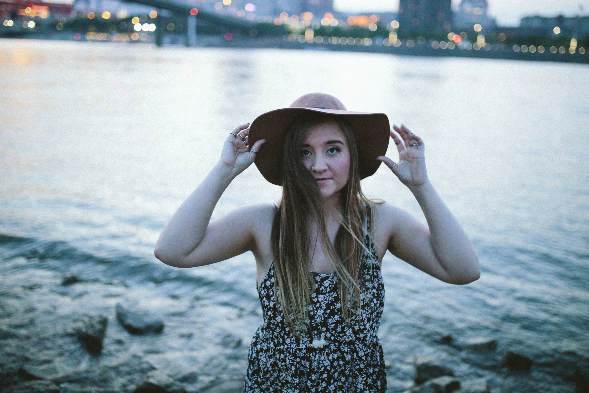 A young woman with long hair wearing a floral dress and a large hat standing near water, with city lights and a bridge in the background during dusk.