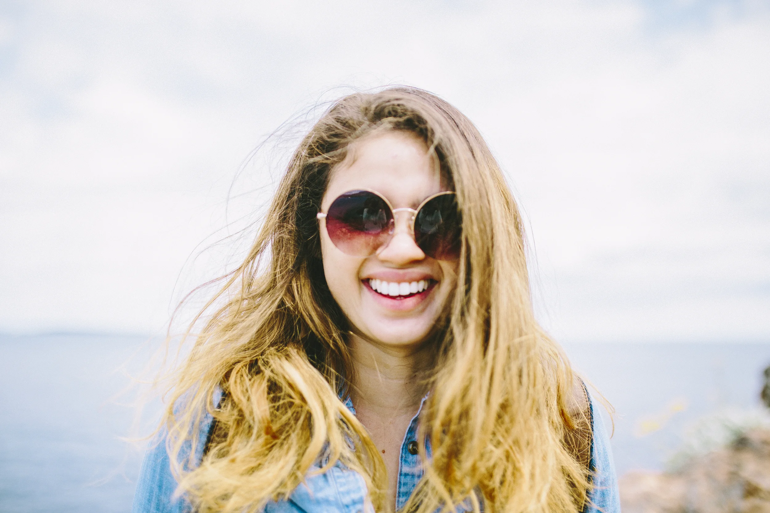 Smiling woman with wavy blonde hair wearing large round sunglasses and a blue shirt outdoors, with a blurred beach and sky in the background.