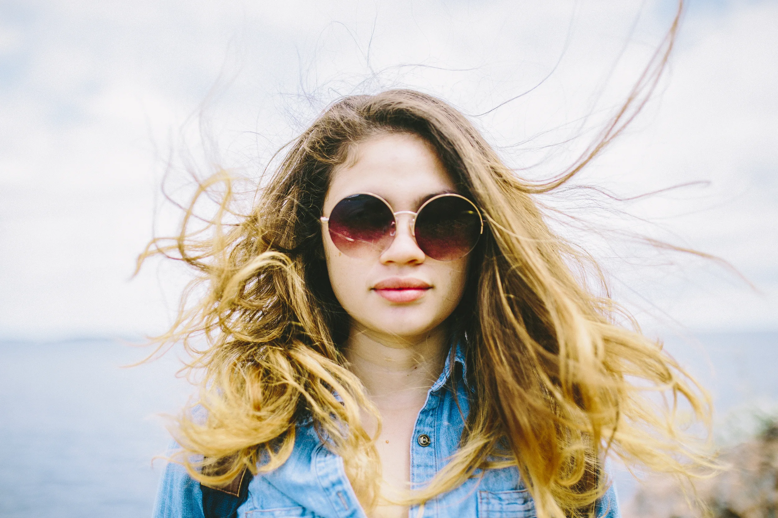 A young woman with wavy, reddish-brown hair wearing sunglasses and a denim jacket outdoors near a body of water.