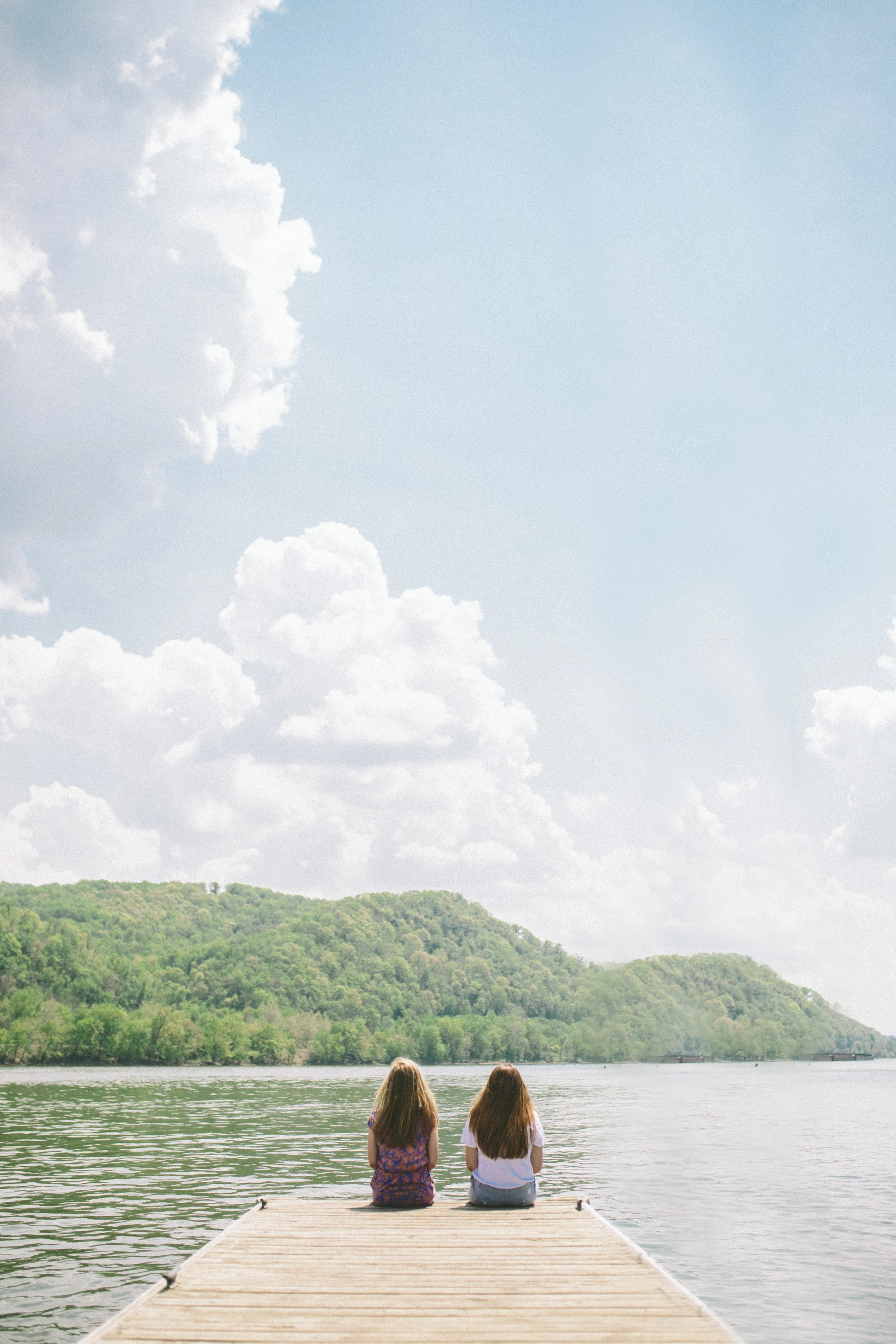 Two girls sitting on a dock by the water, facing a tree-covered hill under a partly cloudy sky.