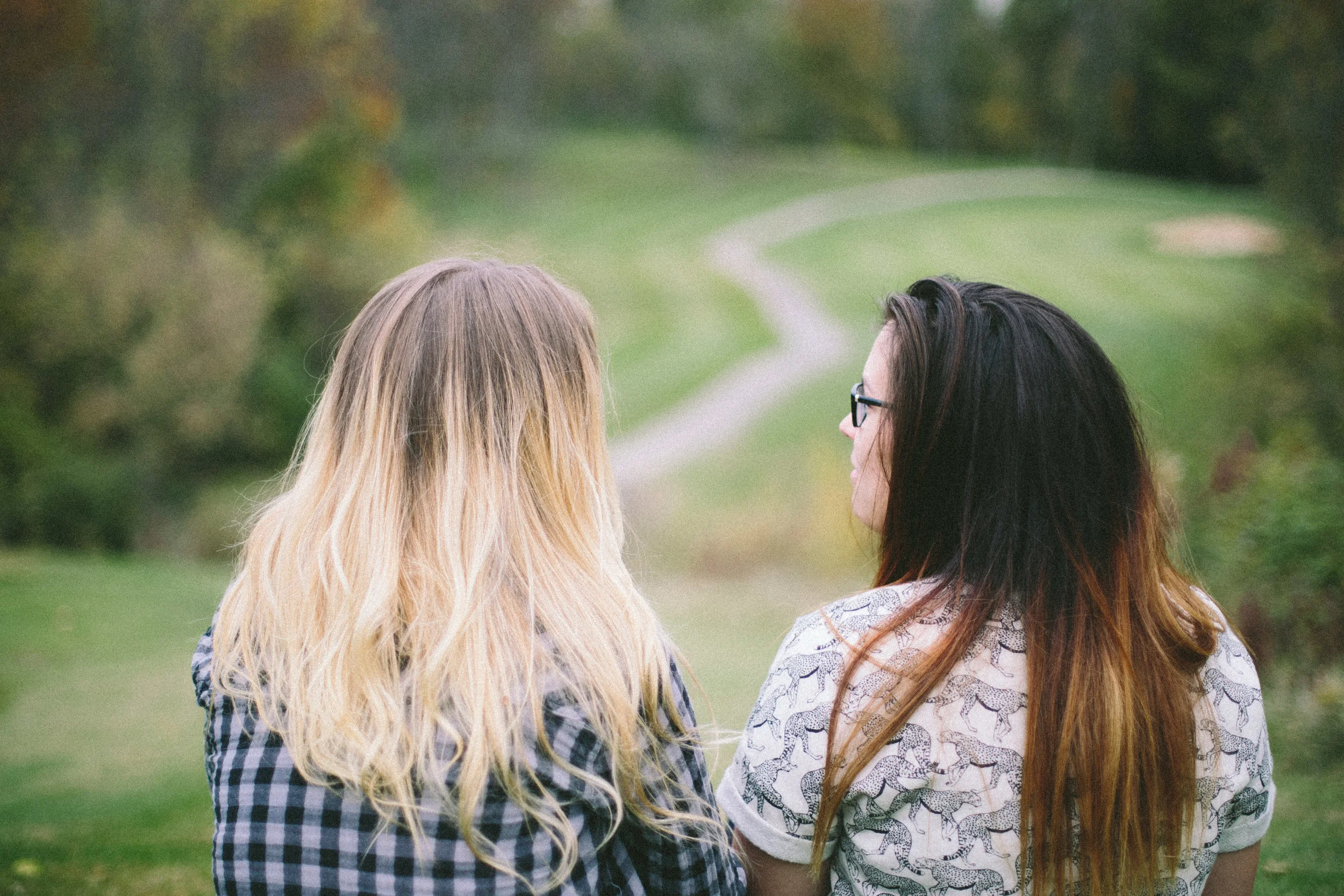 Two women with long hair, one with blonde hair and the other with dark hair, standing outdoors on a grassy path surrounded by trees.