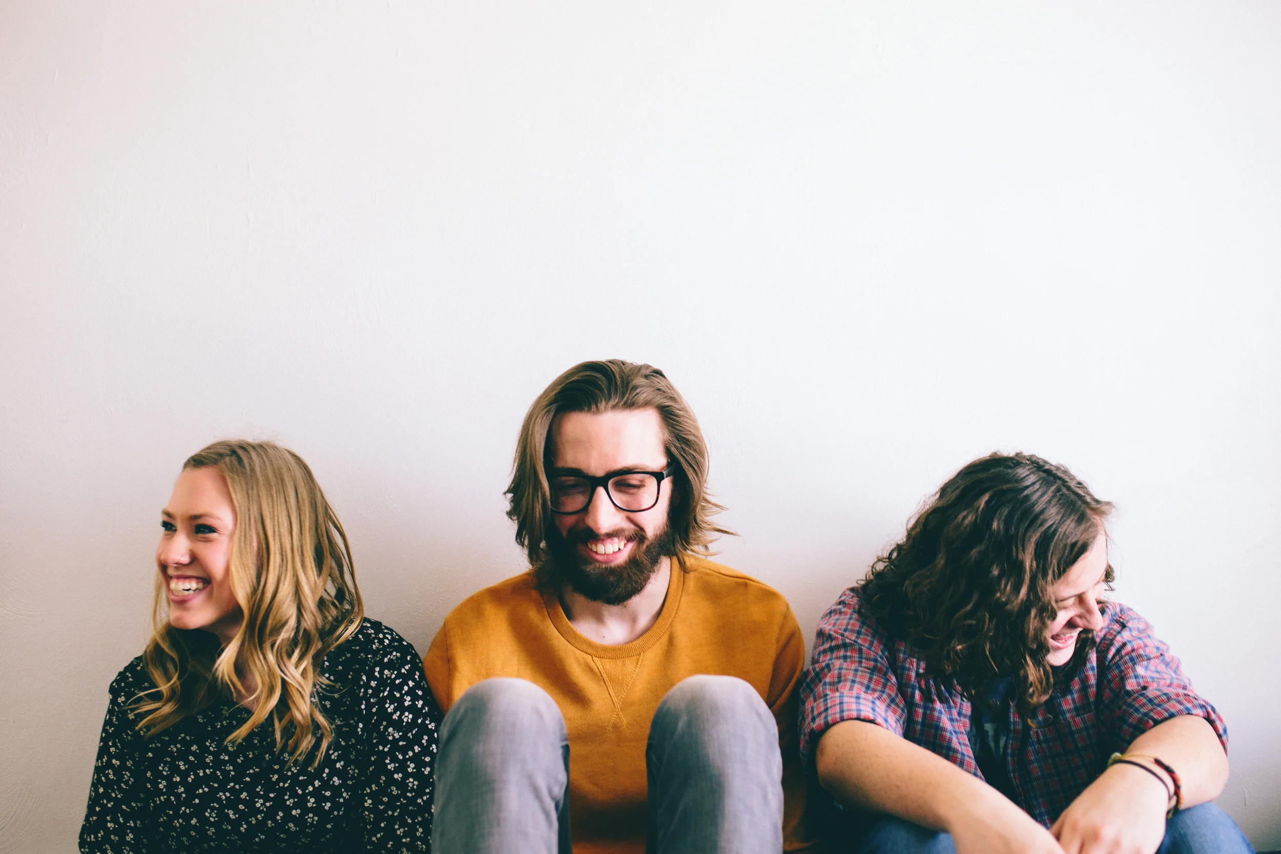 Group of smiling friends sitting against a white wall, engaged in conversation.