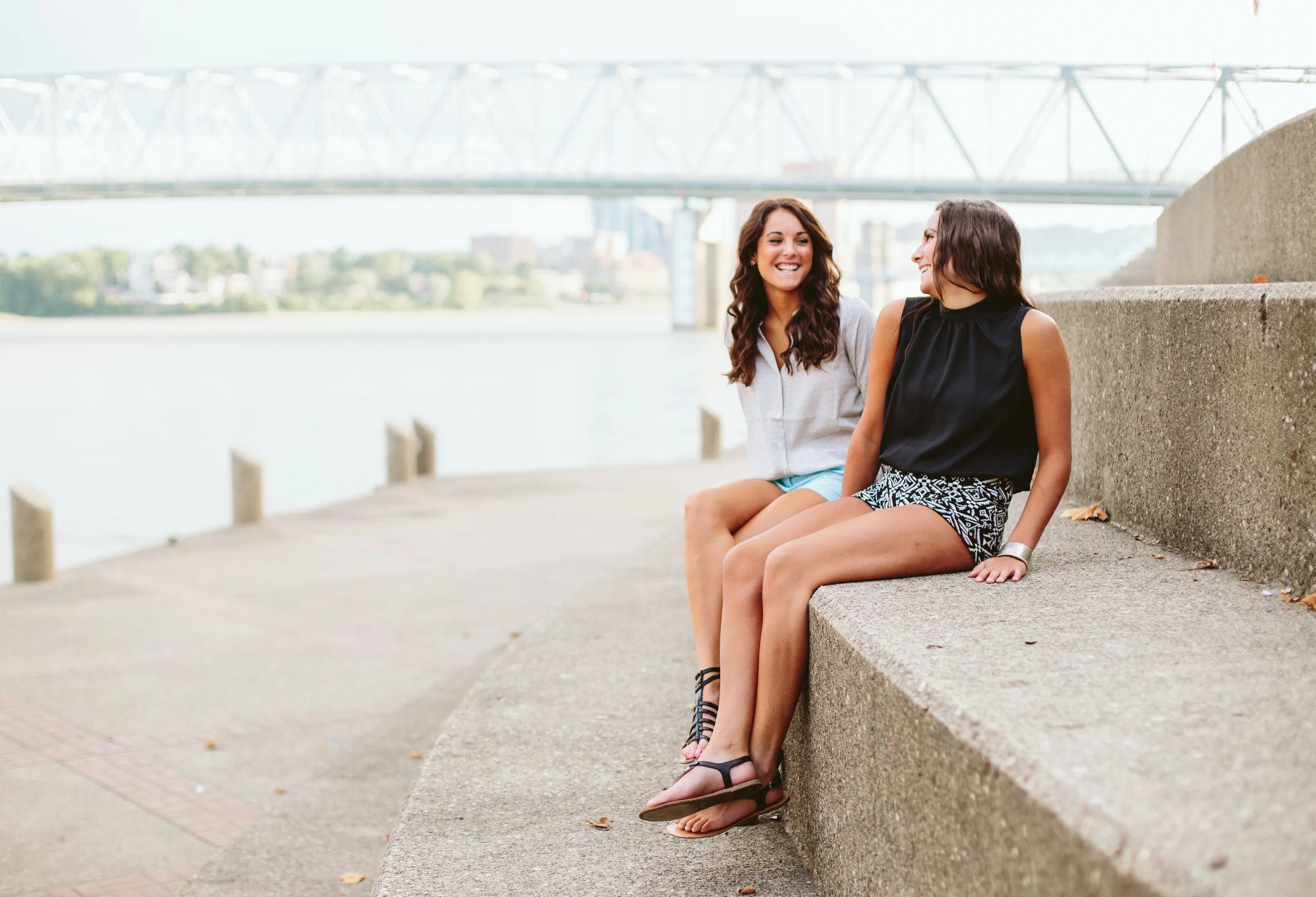 Two women are sitting on a concrete ledge near a body of water, smiling and talking to each other. One woman has long, wavy brown hair and is wearing a light-colored top, while the other has long, dark hair and is wearing a black sleeveless top and p