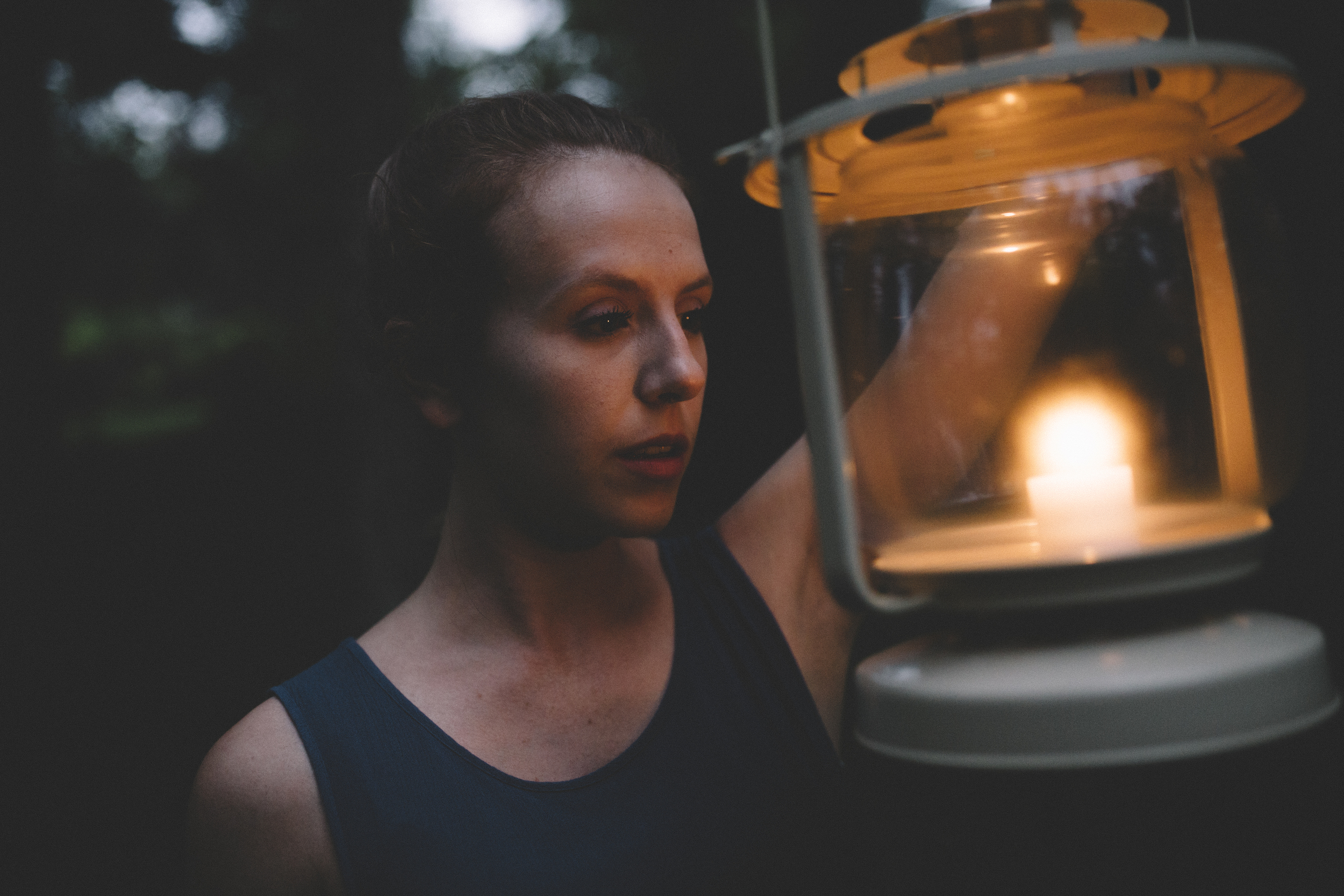 A woman with brown hair wearing a dark sleeveless top, holding an orange lantern with a candle inside, outdoors during dusk or evening.