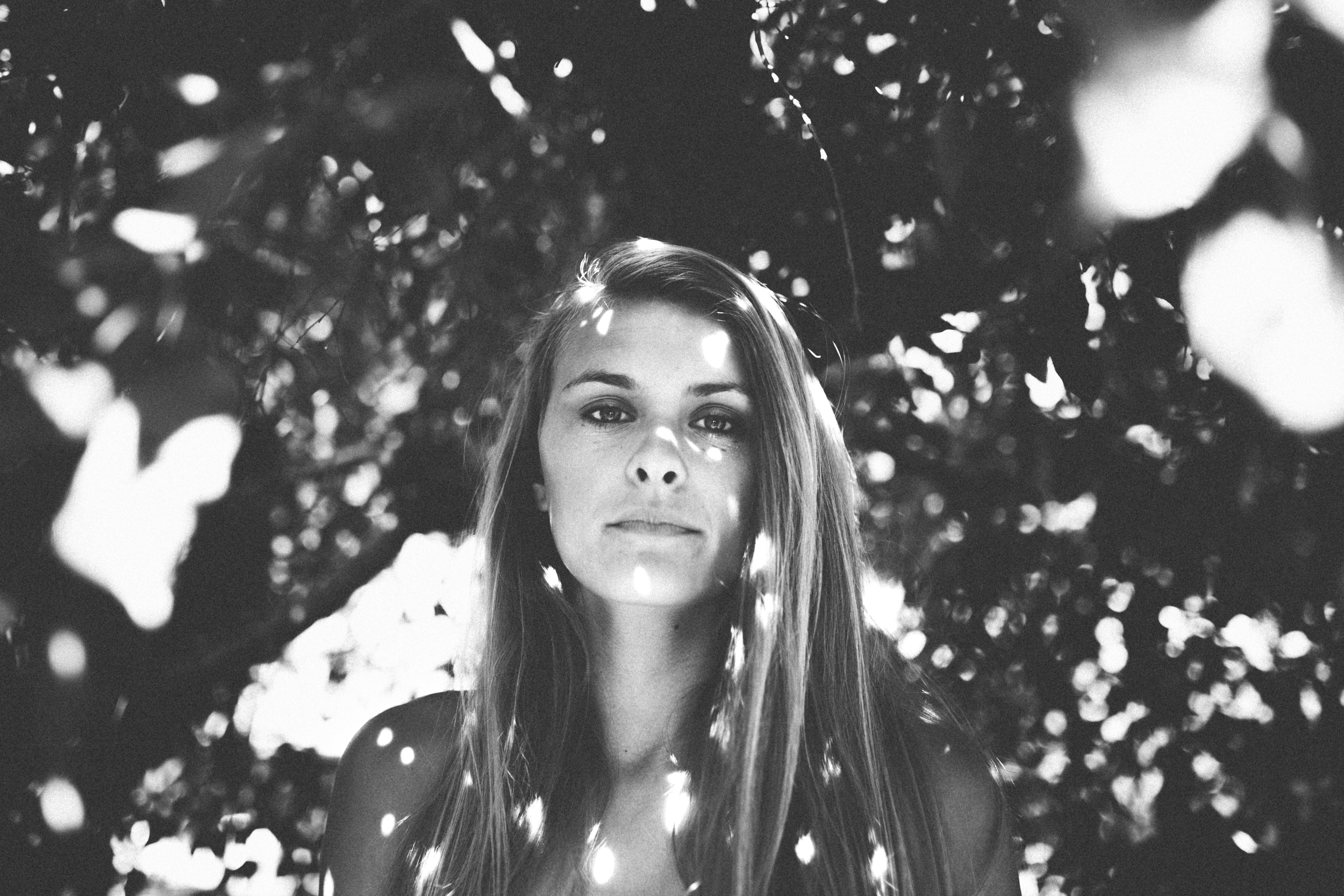 A woman with long hair looking directly at the camera, surrounded by blurry light spots and bokeh effects in black and white.