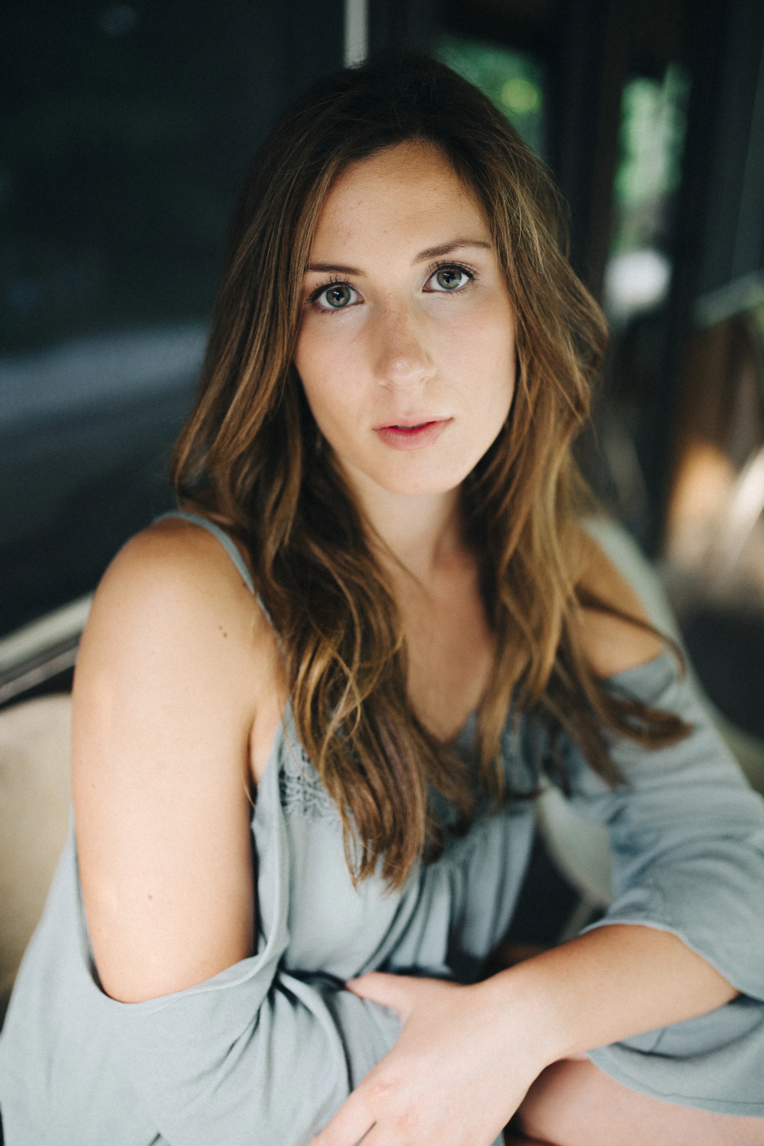 A young woman with long, wavy brown hair and blue eyes sitting indoors with a dark background and greenery outside the window, wearing a light blue off-shoulder top.