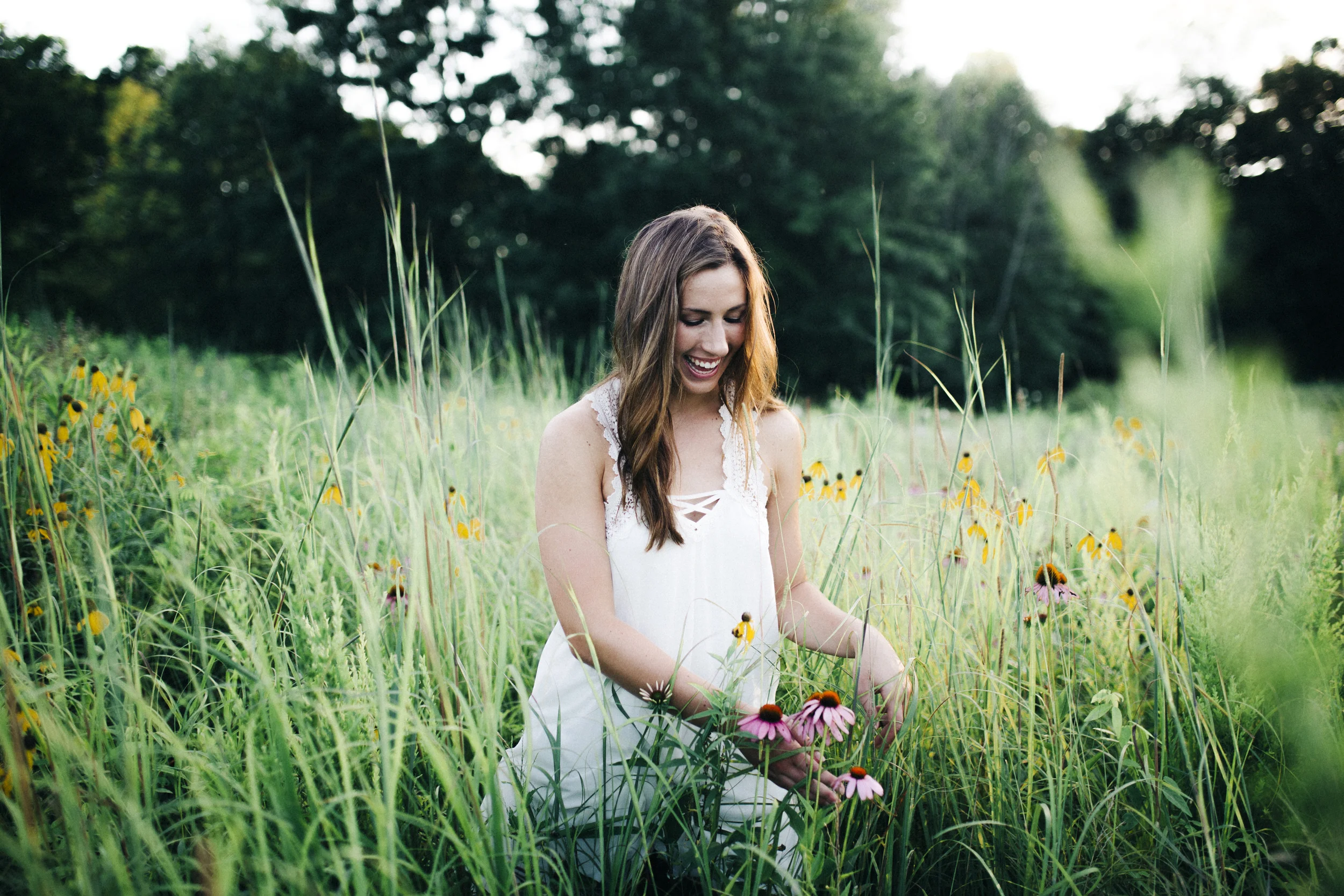 A young woman with long brown hair smiling while sitting in a field of wildflowers surrounded by tall grass and trees in the background.