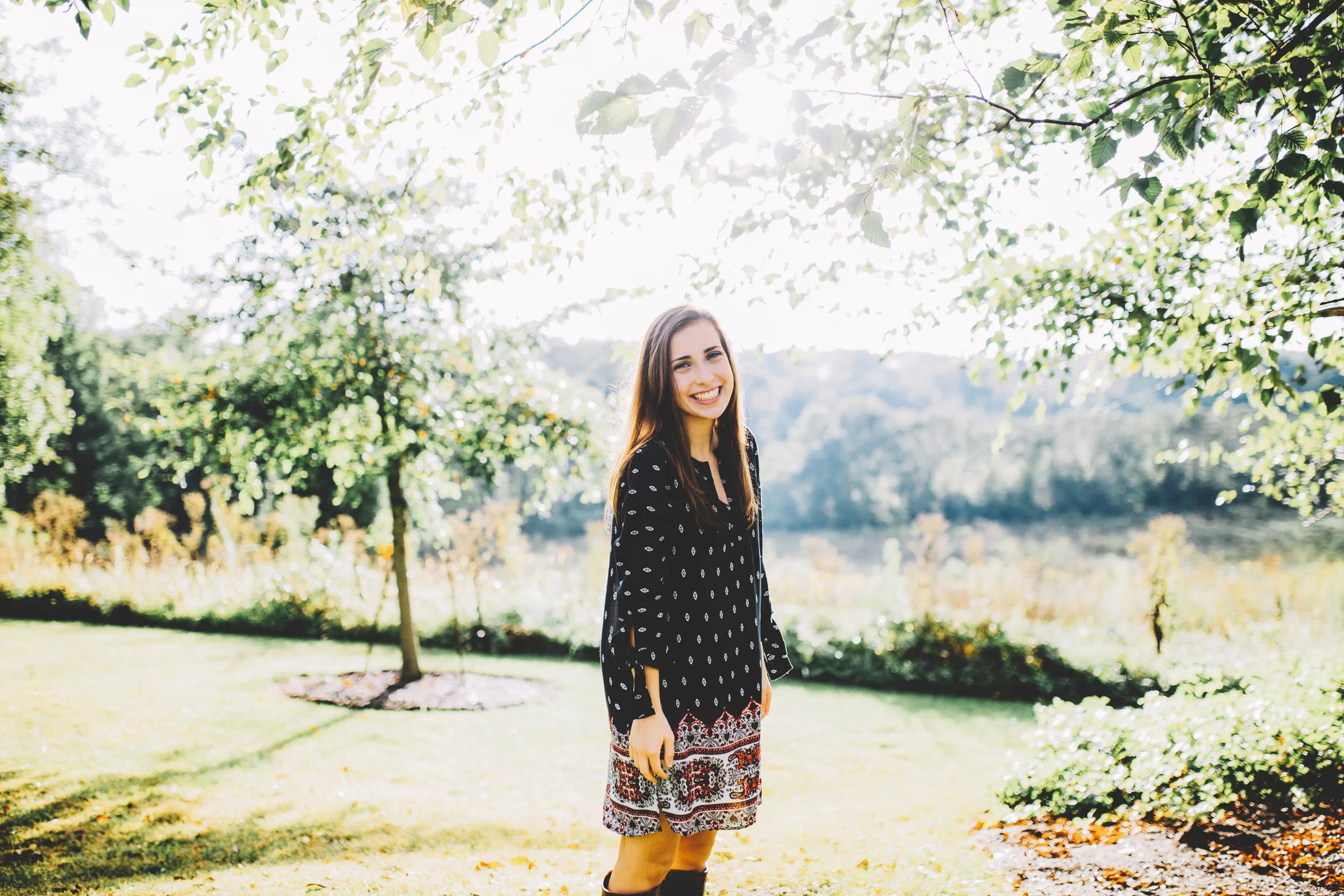 Young woman standing outdoors in a park, smiling, with trees and greenery in the background, sunlight filtering through the leaves.