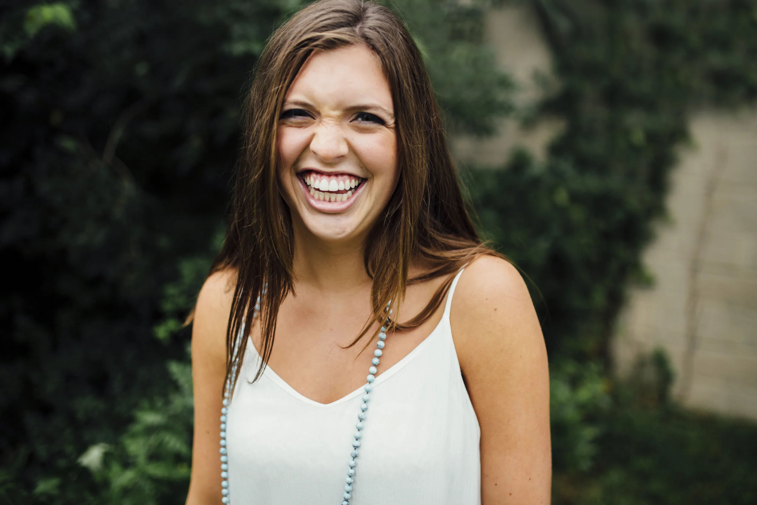 A woman with long brown hair smiling broadly outdoors with greenery in the background.