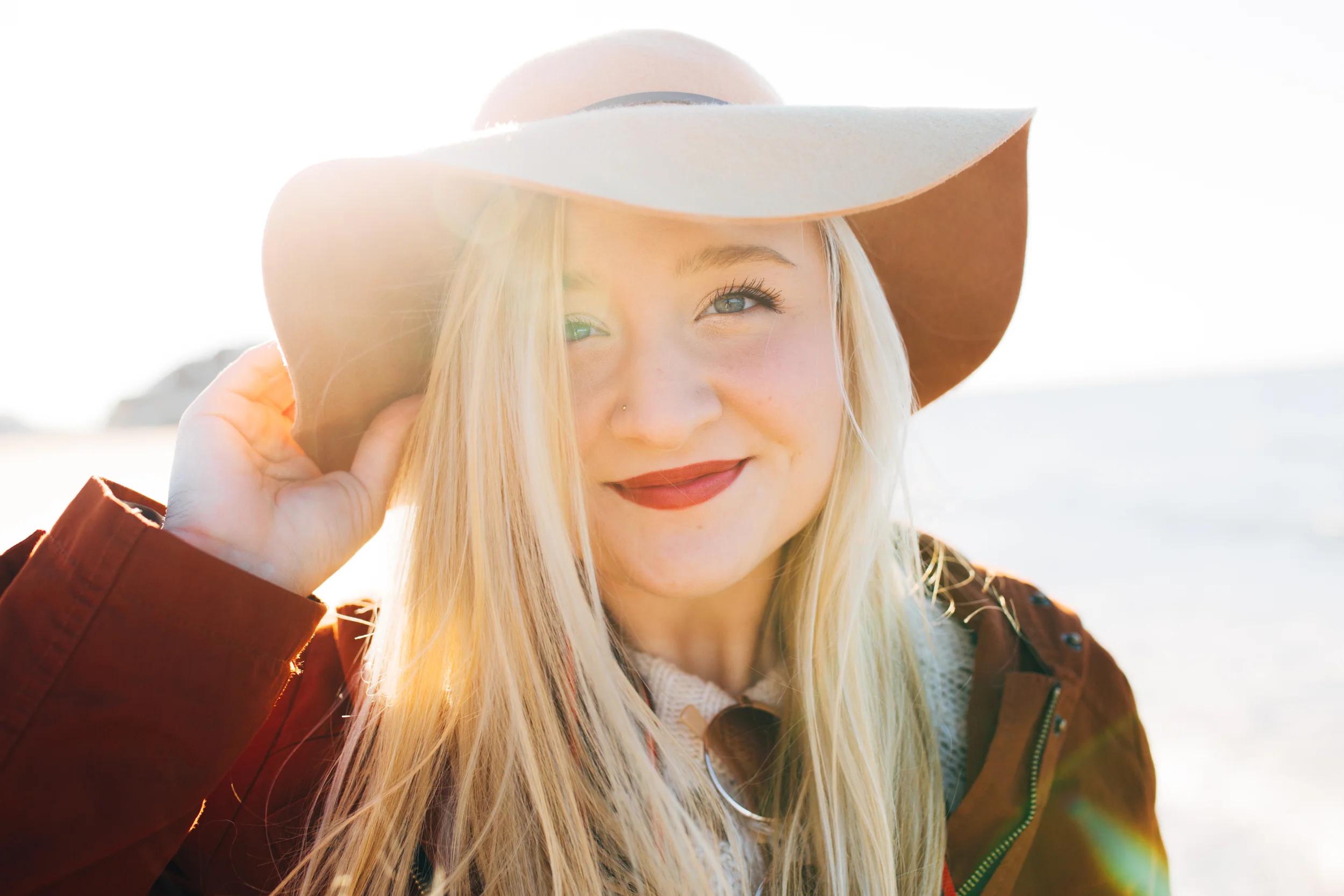 A young woman with long blonde hair and blue eyes smiling, wearing a brown wide-brimmed hat, an orange jacket, and a white sweater, outdoors in bright sunlight.