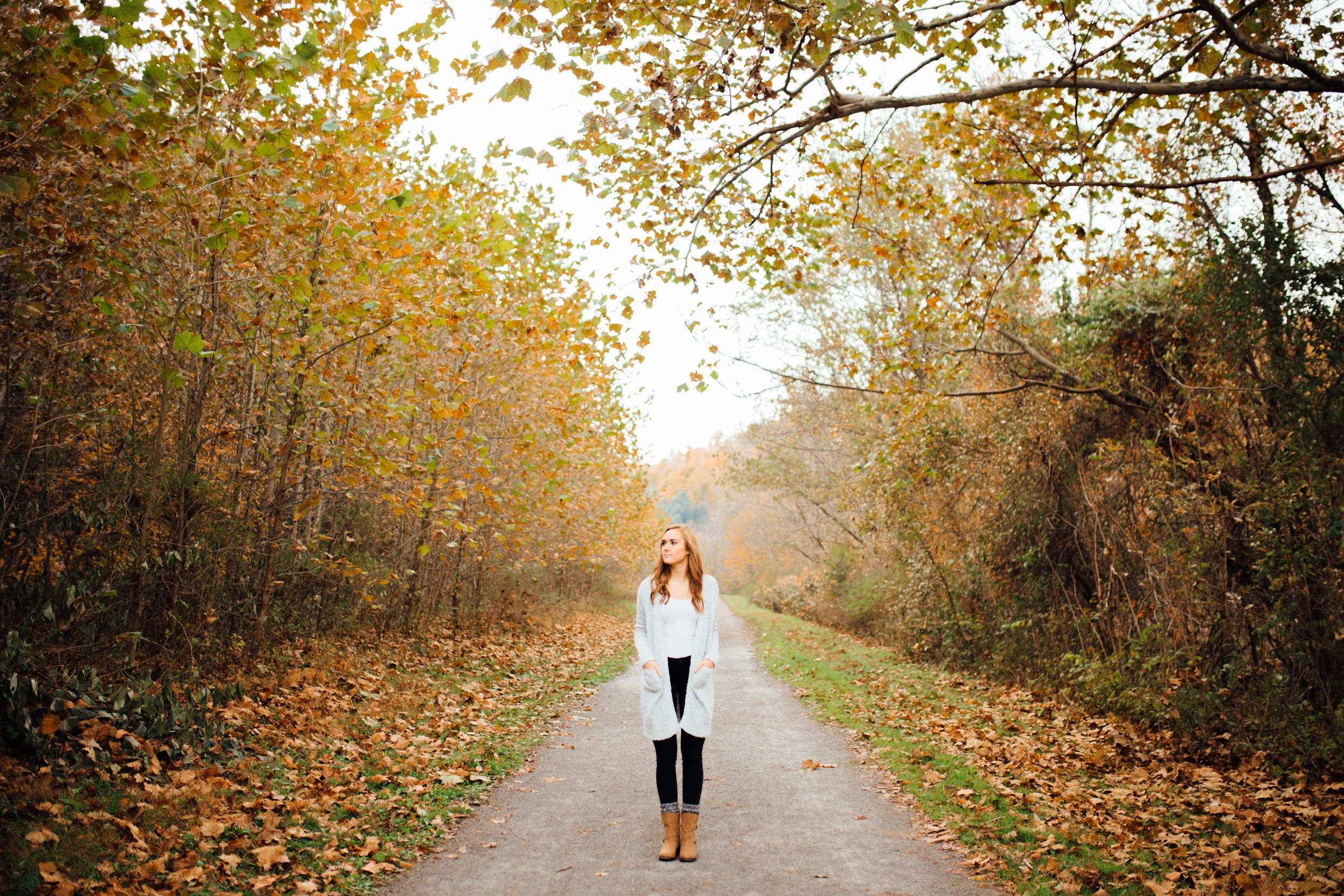 A woman with long hair walking along a leaf-covered path in a wooded area during autumn.