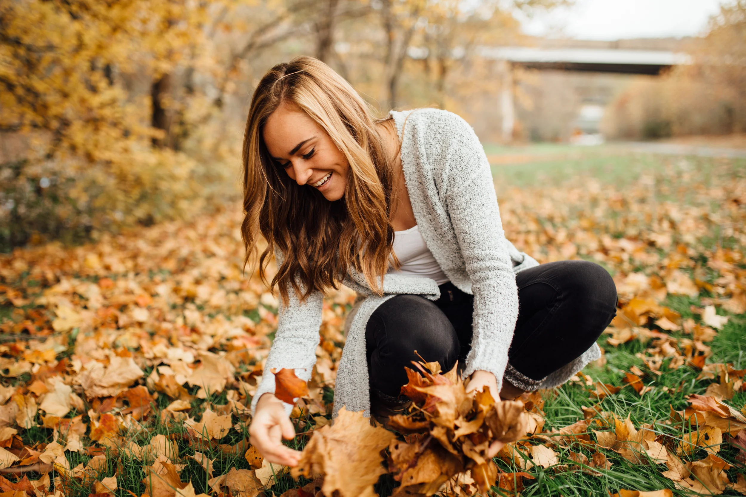 A woman with long brown hair smiling outdoors as she plays with fallen autumn leaves in a park.