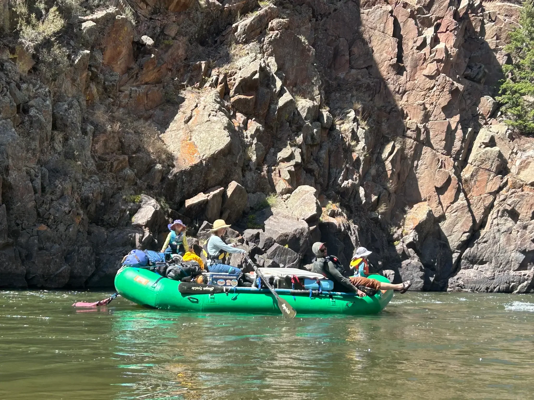 Photo of a whifamily rafting trip: four people—two adults and two children wearing sun hats and life jackets—ride in a green inflatable raft loaded with gear, floating on a calm stretch of greenish river water beneath a tall, rugged rock canyon wall.