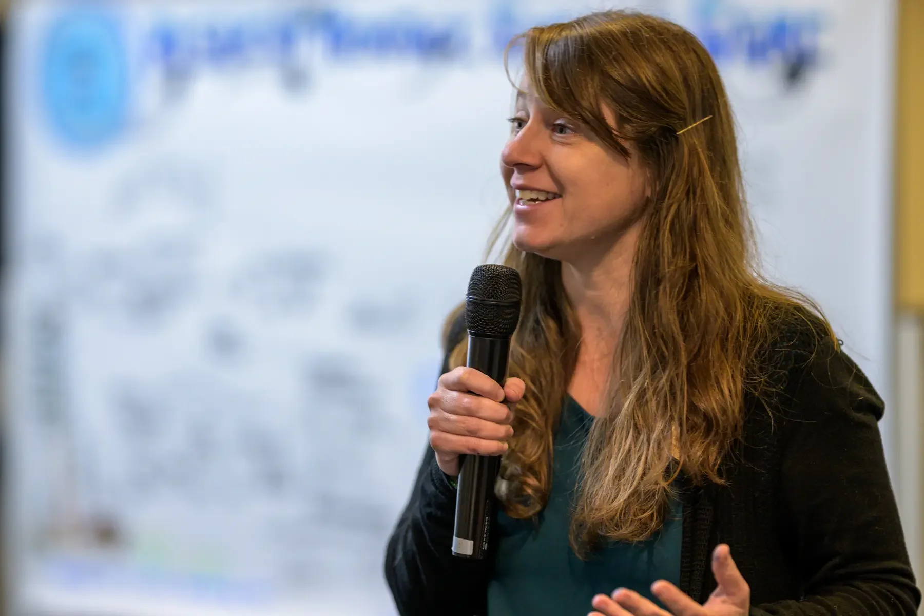 white woman with light brown hair holding a microphone speaking to a group with a graphic recording blurred in the background