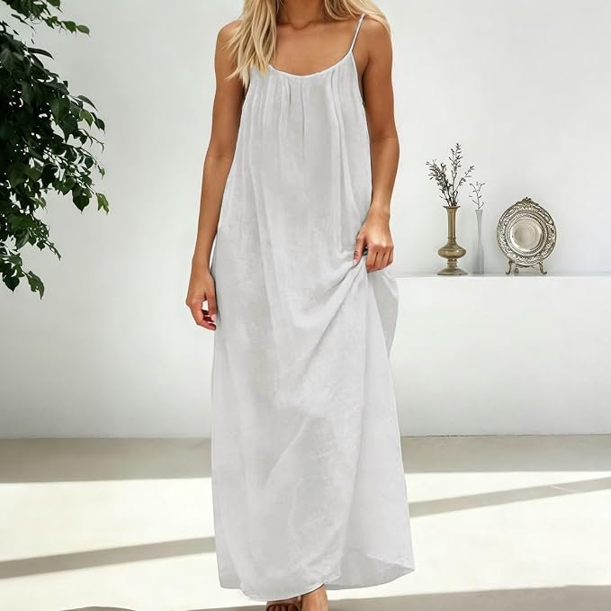 Woman in white maxi dress standing indoors with a white wall, potted plant, and decorative objects in the background.