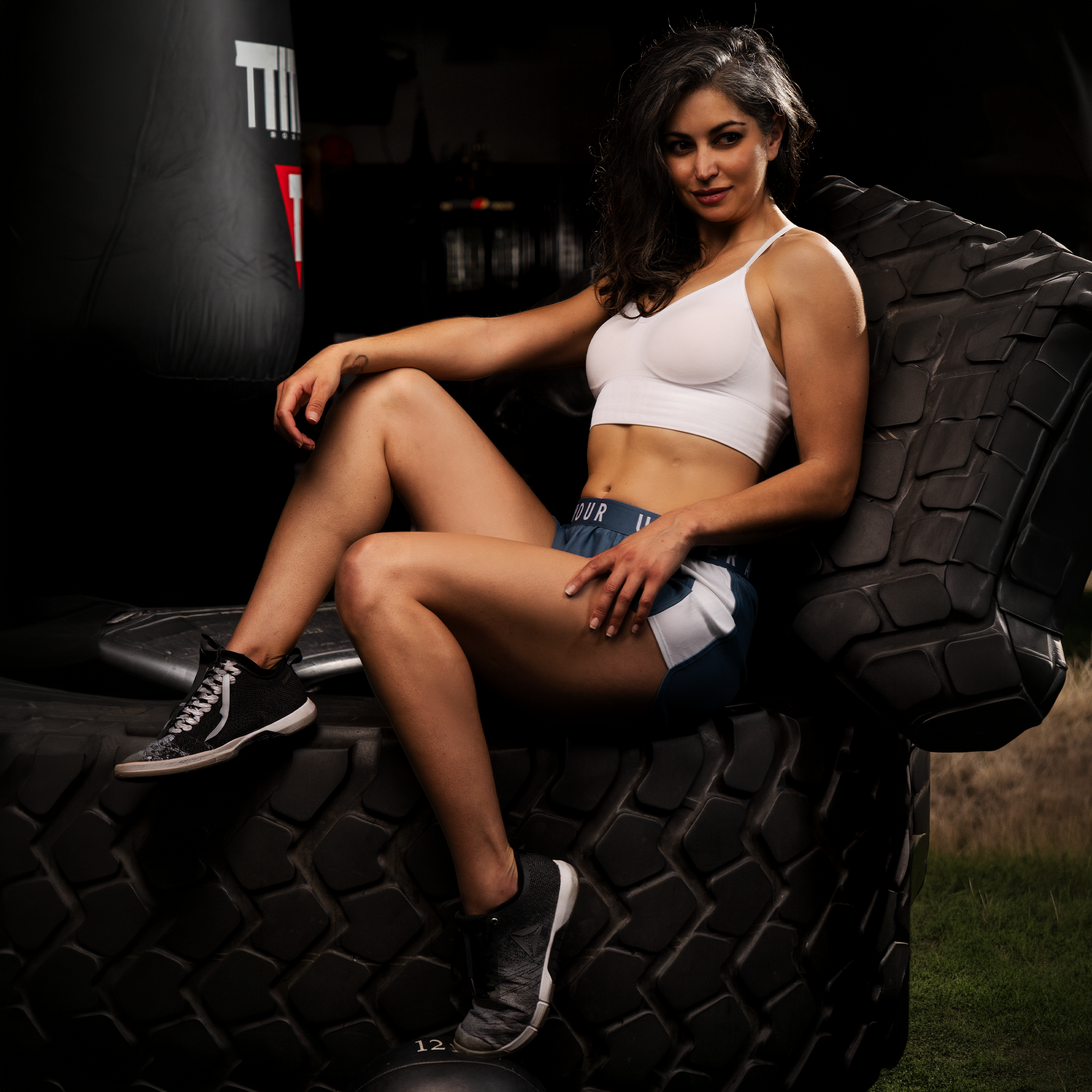 A woman sitting on a large black tire in a dark gym with boxing equipment, wearing a white sports bra, shorts, and black sneakers.