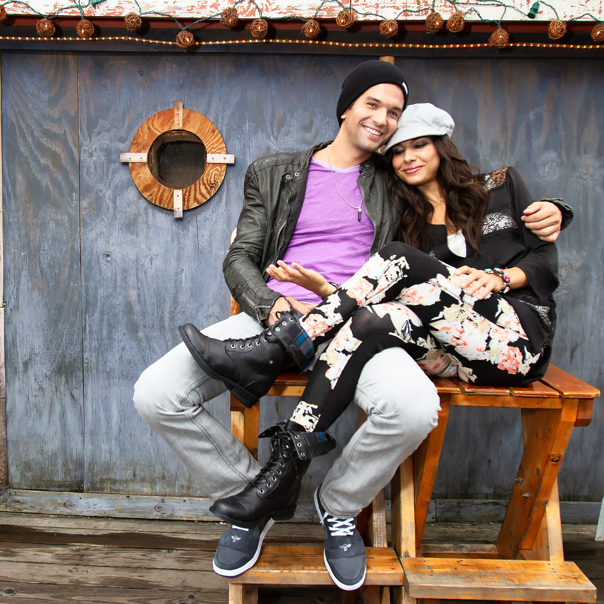 Young man and woman sitting on a wooden bench with a blue wall behind them, smiling and embracing. The man is wearing a black beanie, purple shirt, and leather jacket, and the woman is wearing a gray hat and a black lace top. There are decorative str