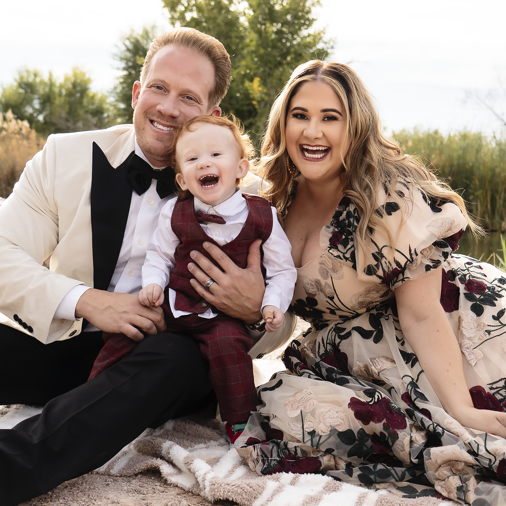 A happy family outdoors, with a man in a white tuxedo, a woman in a floral dress, and a young boy in a red plaid vest, smiling and posing for a photo during daytime.