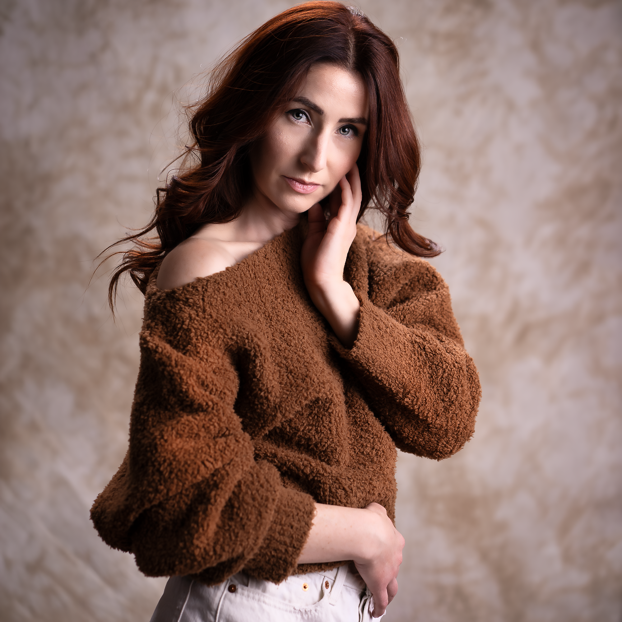 A woman with long, curly reddish brown hair and blue eyes, wearing a brown fuzzy sweater and light-colored pants, stands against a beige textured background. She has a gentle expression and her hand is touching her face.