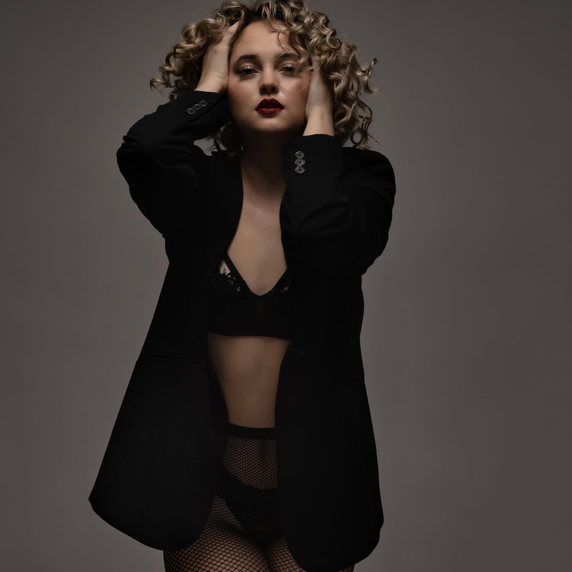 A woman with blonde curly hair and gold eyeshadow poses with her hands on her head, wearing a black blazer, lace bodysuit, and fishnet tights against a plain gray background.