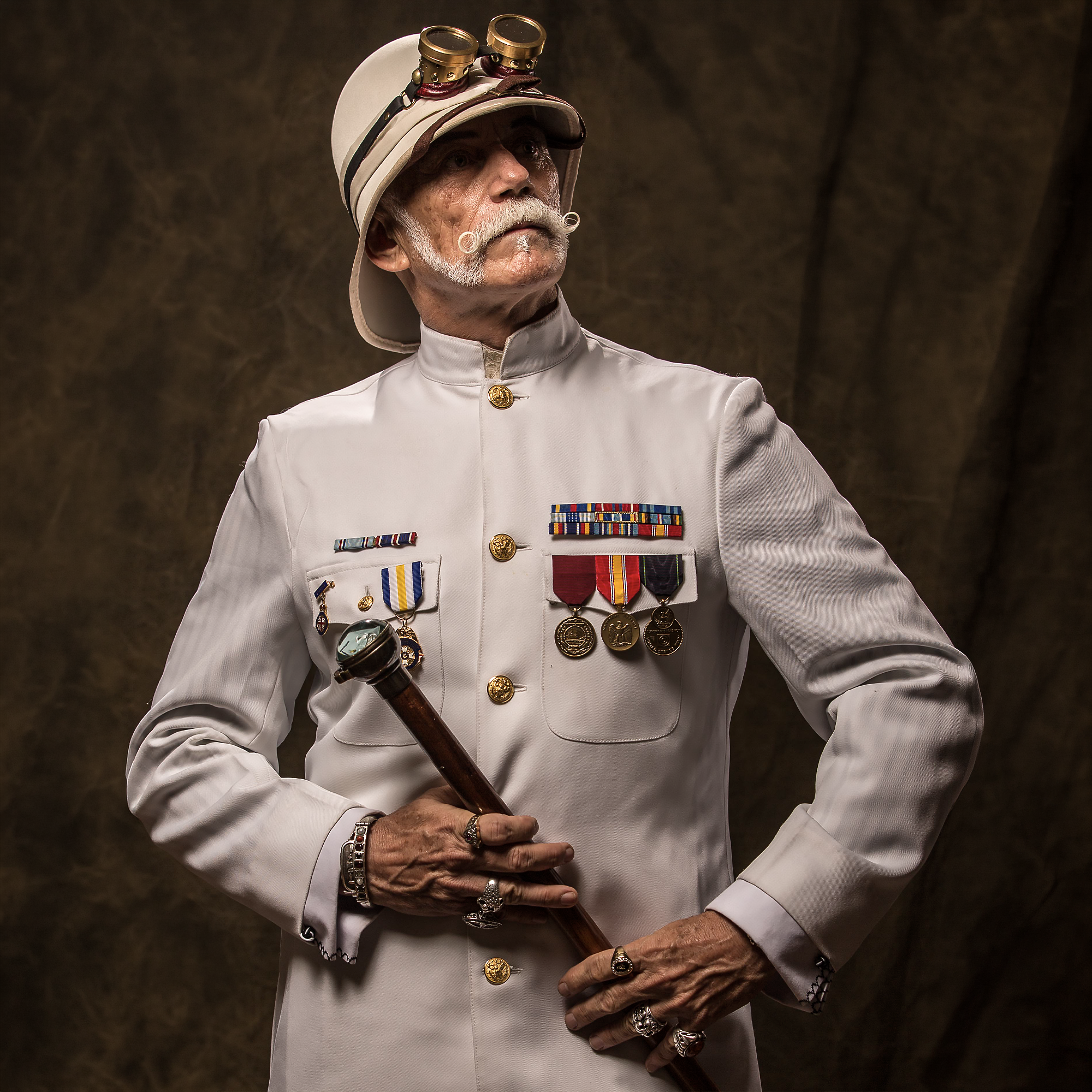 An elderly man dressed in a formal military uniform with numerous medals and ribbons, wearing a helmet with goggles, holding a ceremonial sword, and wearing various jewelry.