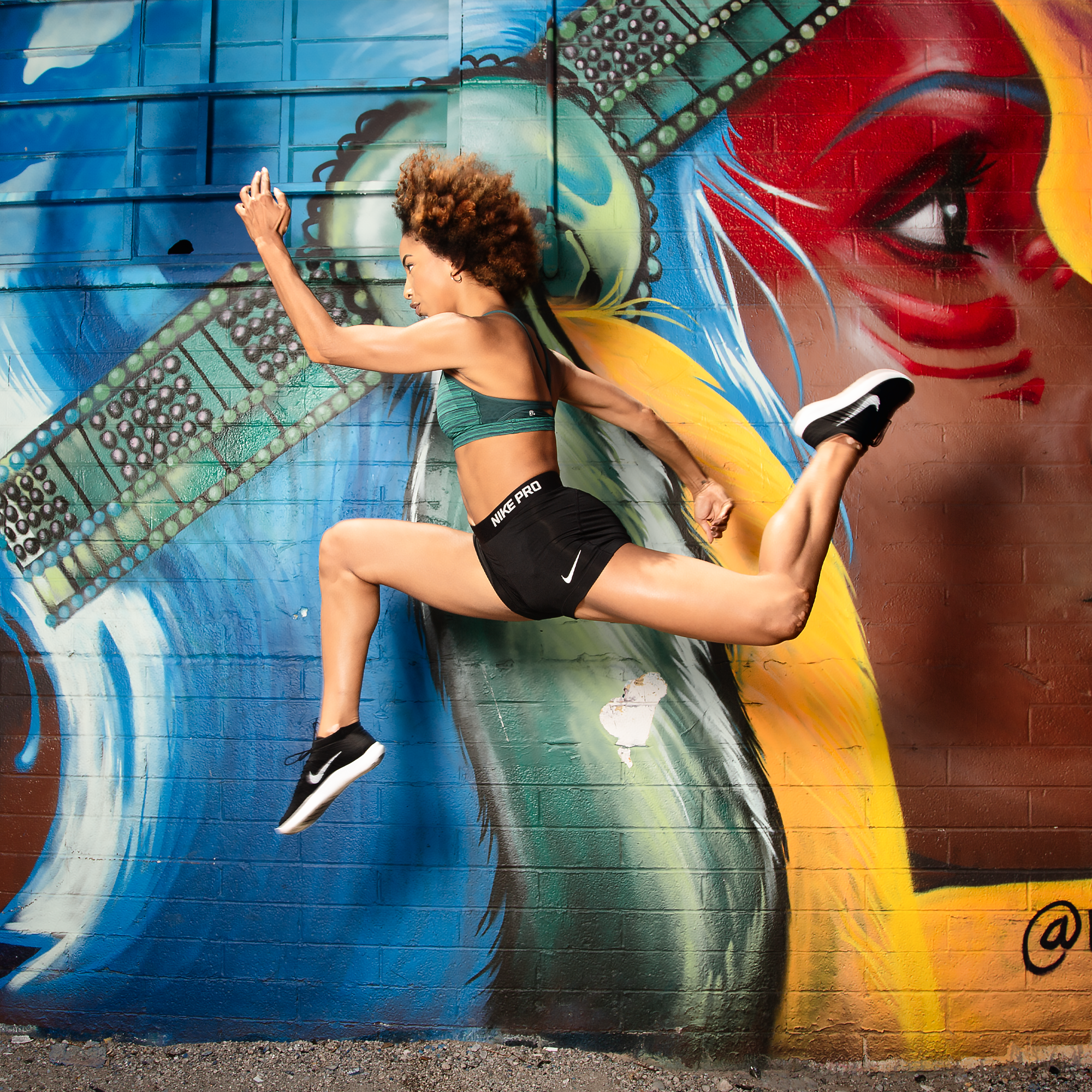 A woman in workout attire, including a green sports bra and black Nike Pro shorts, performing a plank against a colorful graffiti wall featuring a large butterfly and a woman's face with red lips.