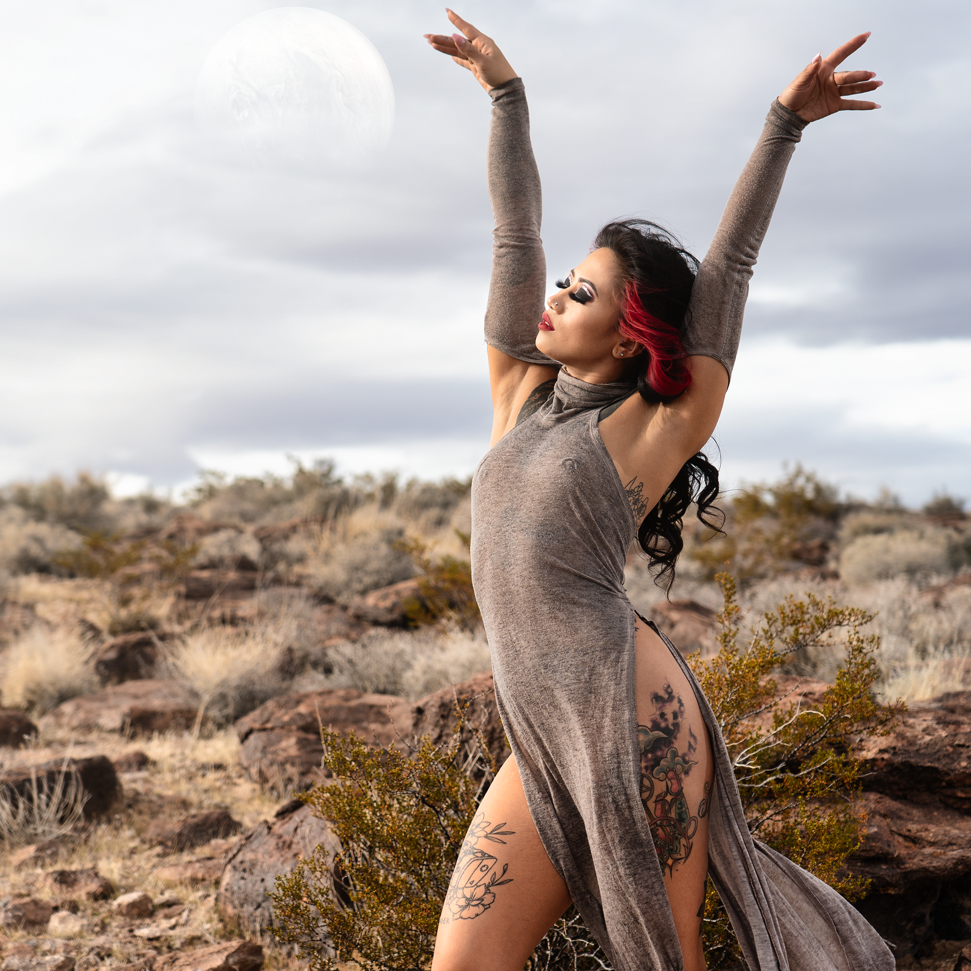 A woman with dark hair and red streaks, tattoos on her arm and side, wearing a sleeveless gray dress and gray long sleeves, is outdoors on a cloudy day with her arms raised and hands reaching towards the sky, with a large moon visible in the backgrou