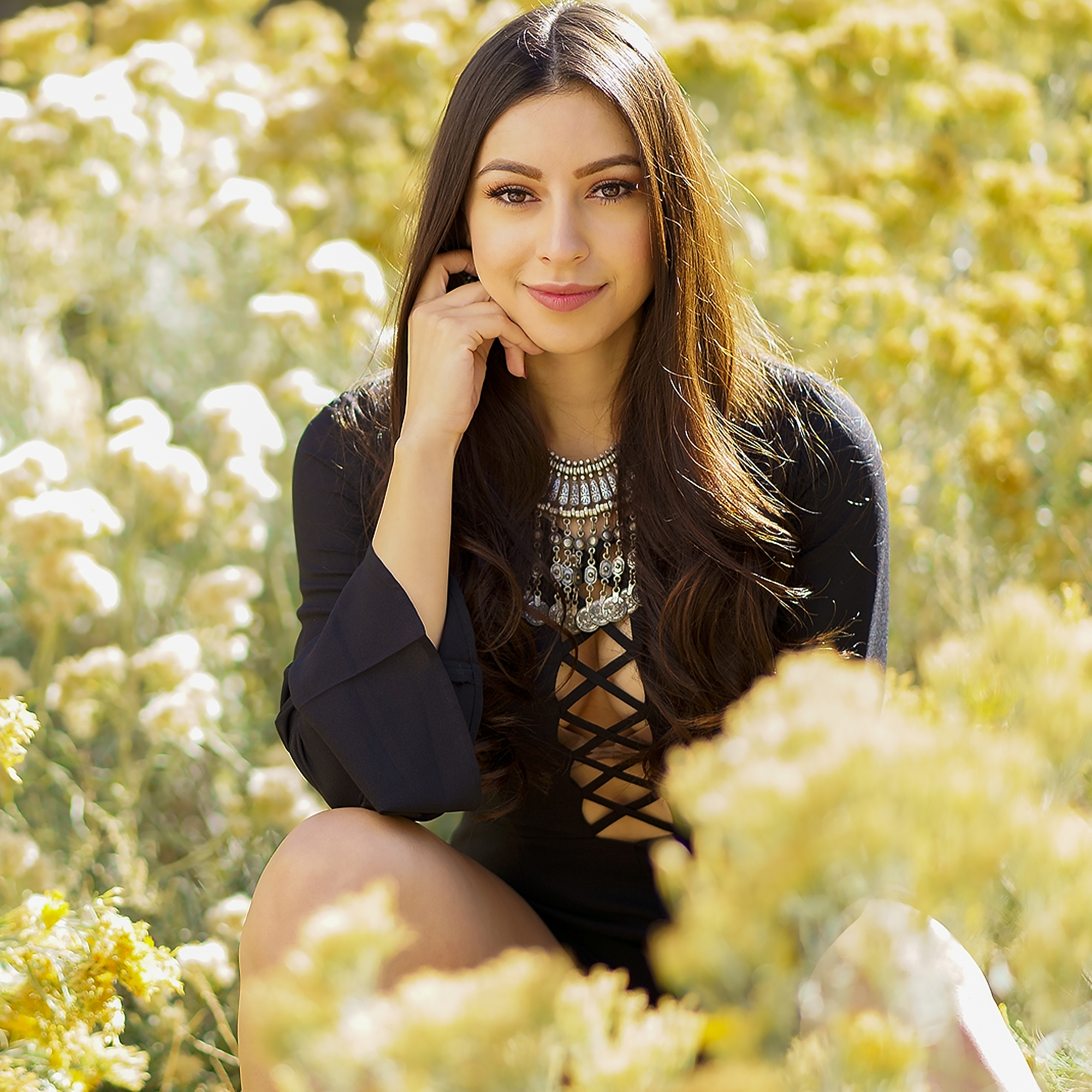 A young woman with long dark hair and makeup, smiling with one hand resting near her face, wearing a black lace-up dress and layered necklaces, outdoors among yellow and white flowers with blurred trees in the background.