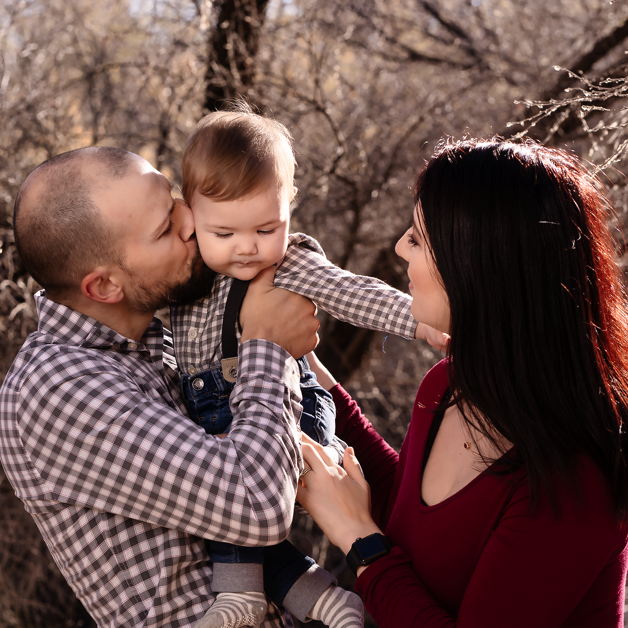 A family of three outdoors, a man, woman, and young child, enjoying a moment together during daylight. The man kisses the child's cheek, and the woman looks at the child. They are dressed casually, with trees or bushes with bare branches in the backg