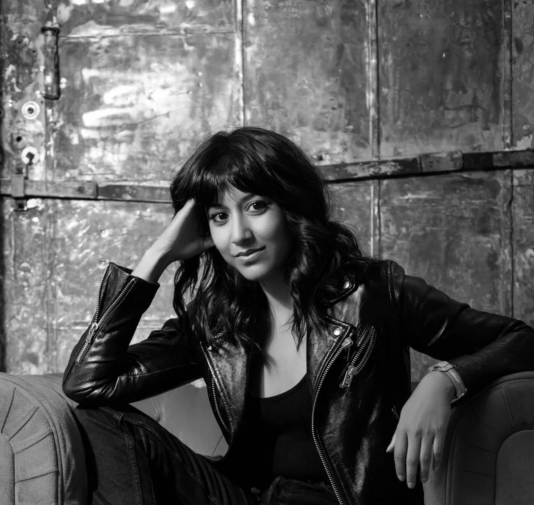 A black and white portrait of a woman with wavy hair, wearing a leather jacket and sitting on a couch with her arm resting on the back, in front of a textured wall.