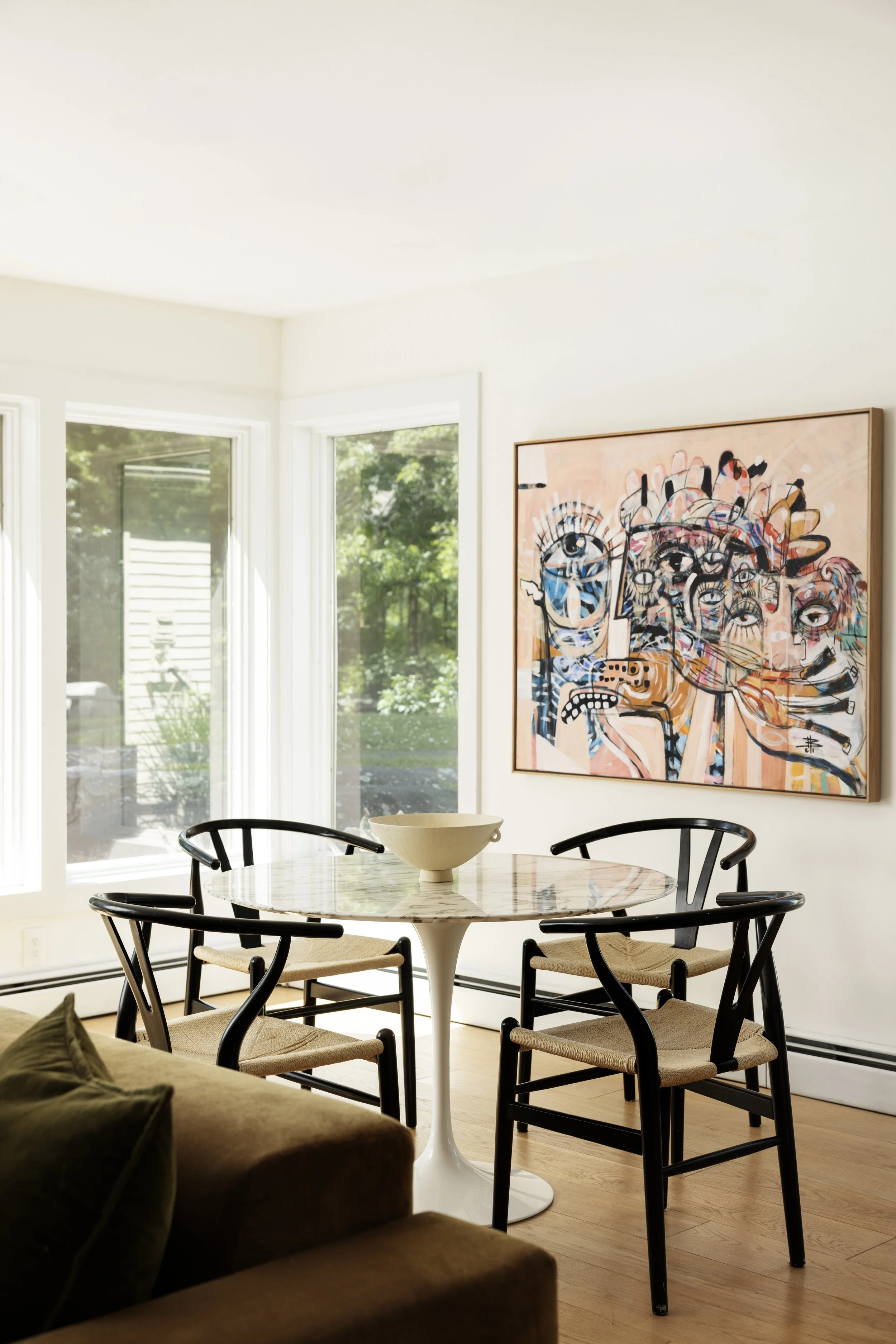 A dining area with a round marble table and four black chairs with beige seats, a large abstract painting of multiple faces on the wall, and natural light coming through three windows, with green trees outside.