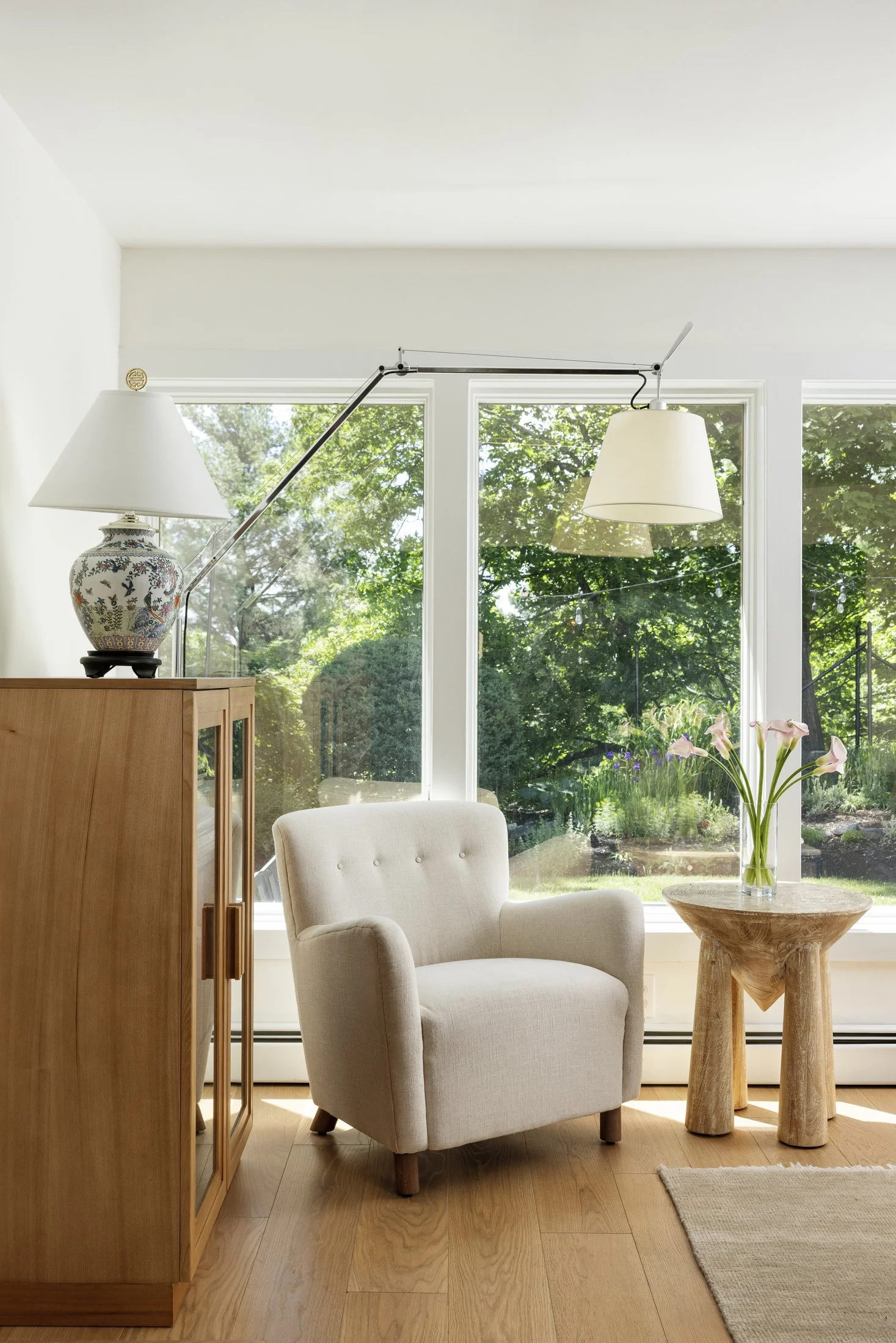 Living room with a large window, a white armchair, a wooden sideboard with a lamp, a small wooden table with a vase of pink lilies, and a hanging white lampshade.