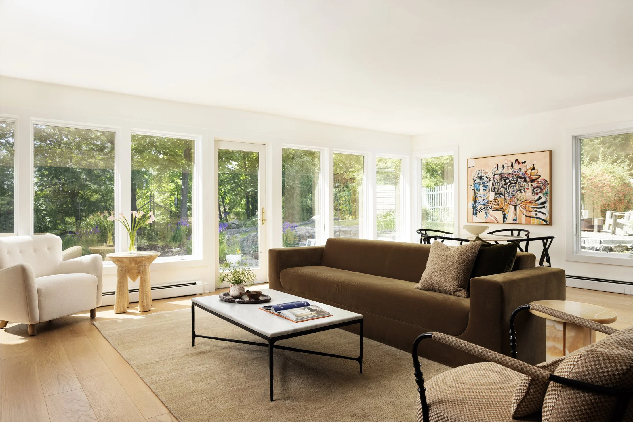 Bright living room with large windows overlooking greenery, featuring a brown sofa, a beige armchair, a black coffee table with magazines, and colorful modern artwork on the wall.