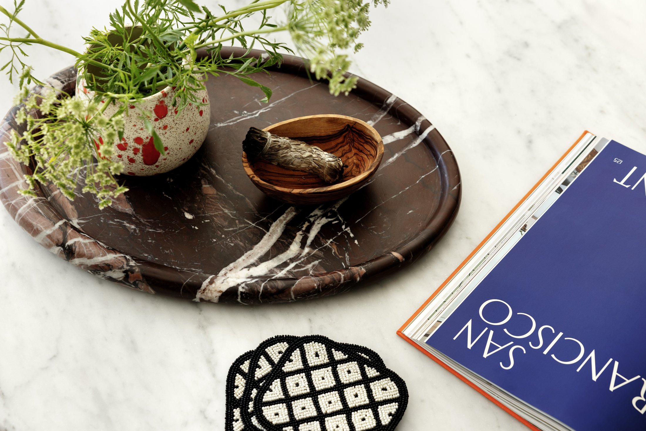 A marble tray with a plant in a speckled pot, a wooden bowl with a cigar band, and a stack of magazines on a white surface.