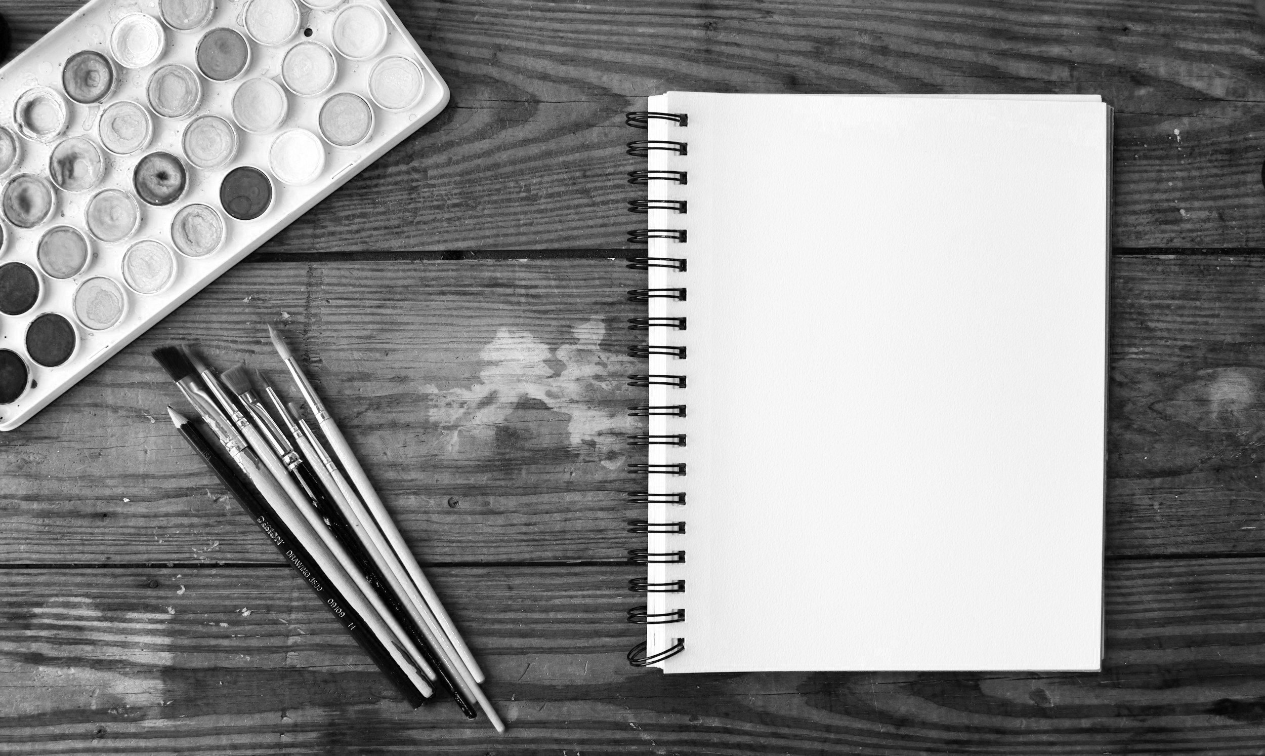 A wooden desk with a palette of watercolor paints, four pens, and an open spiral notebook with blank pages.