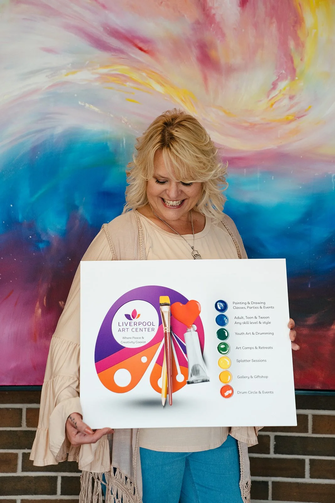 A smiling woman holding a colorful sign for Liverpool Art Center with painting supplies attached, standing in front of a vibrant, abstract wall.