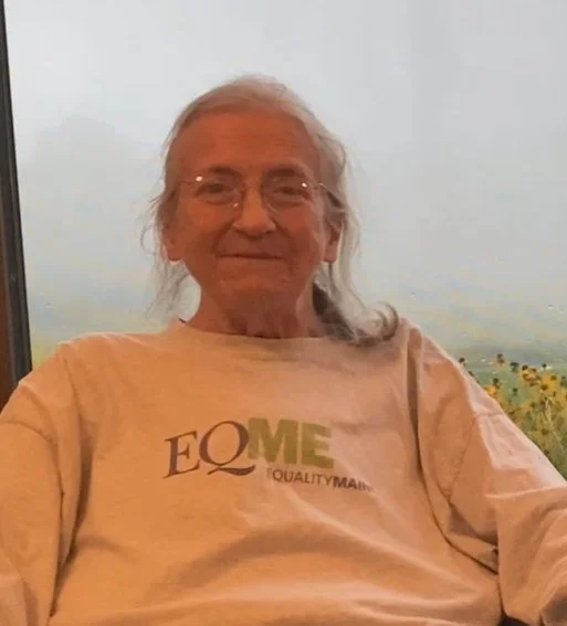 An elderly woman with gray hair and glasses, wearing a beige shirt with the word 'EQUQME' printed on it, sitting indoors with a window showing a cloudy sky and a landscape of greenery and flowers in the background.